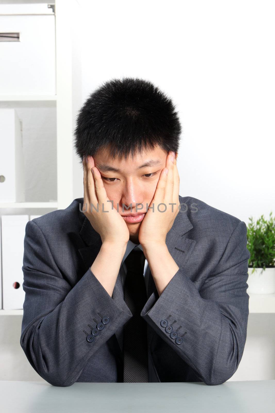 Despondent Asian businessman sitting at his desk with his chin resting in his hands staring morosely at the desktop