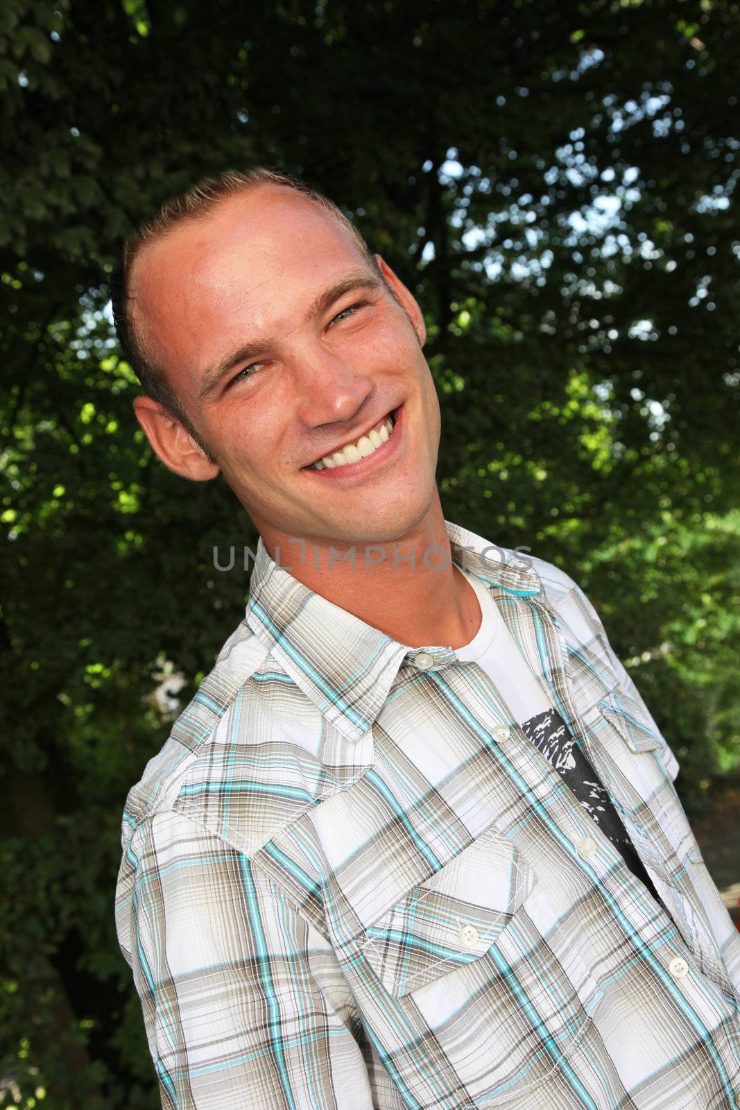 Smiling sexy handsome young man in a casual shirt standing outdoors in front of greenery