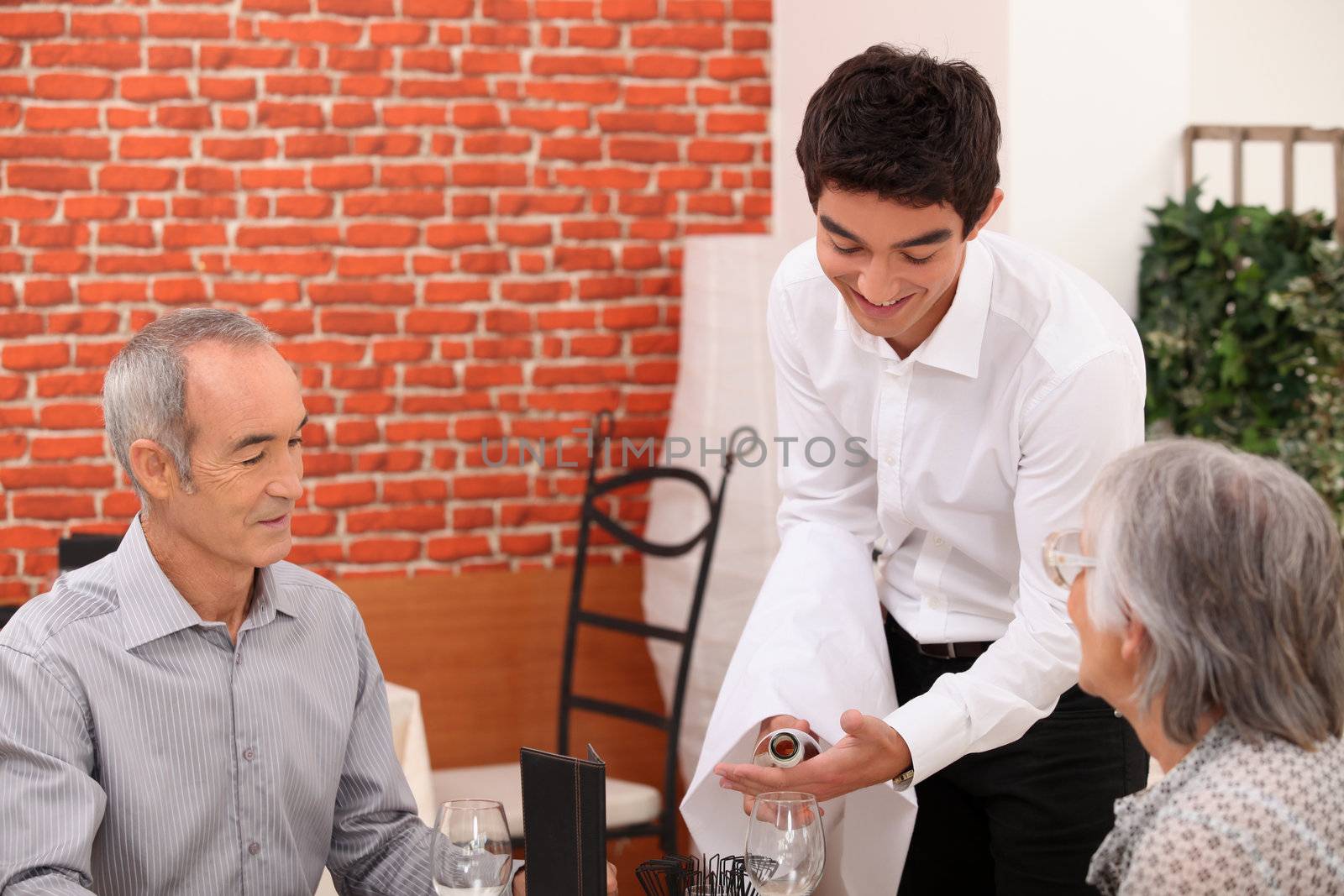 Young waiter pouring wine for an older couple by phovoir