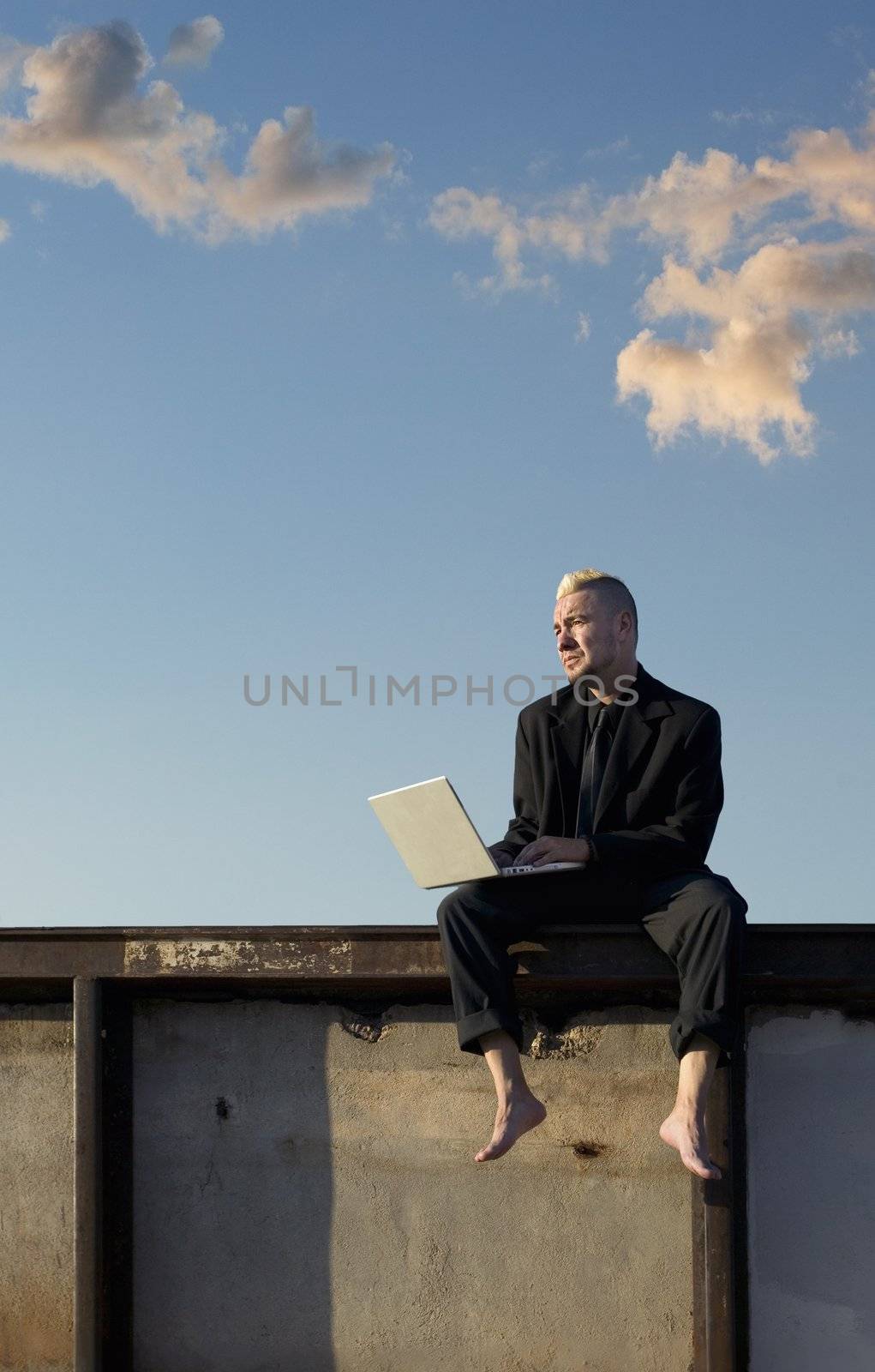 Barefoot businessman with a punk haircut works on his laptop computer sitting on a wall.