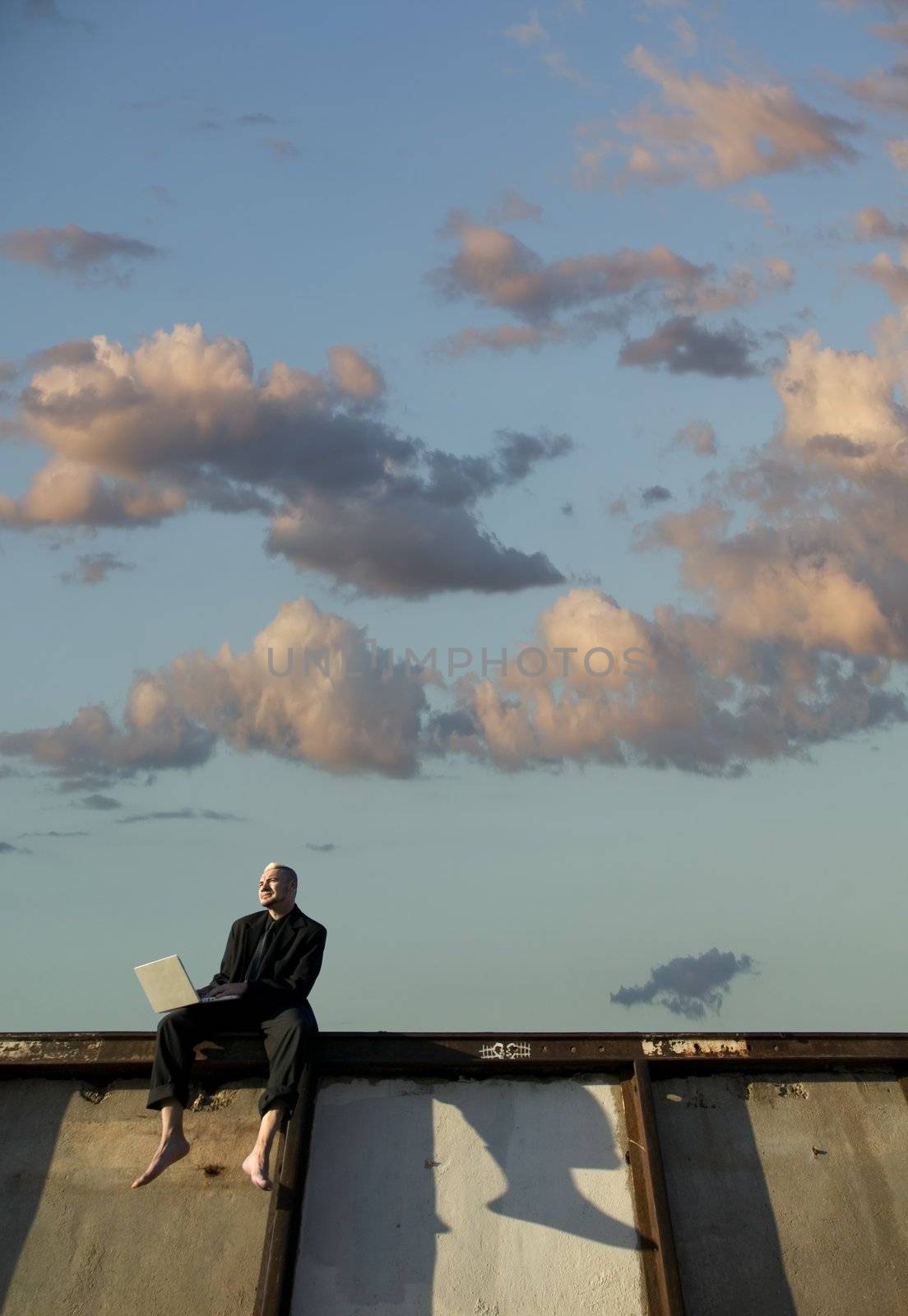 Barefoot businessman with a punk haircut works on his laptop computer sitting on a wall.