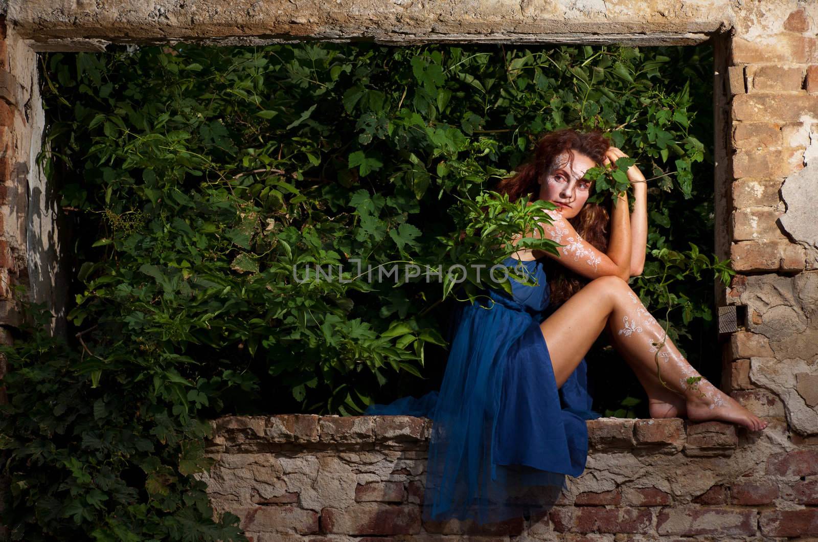 Wild woman with white body henna tattoos posing on gained by bushes window of abandoned building