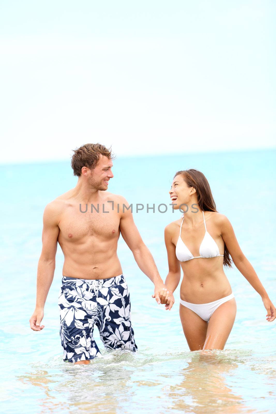 Couple on beach walking in water holding hands having fun during summer holidays travel vacation. Young happy joyful interracial couple, Caucasian man, Asian woman together outside.