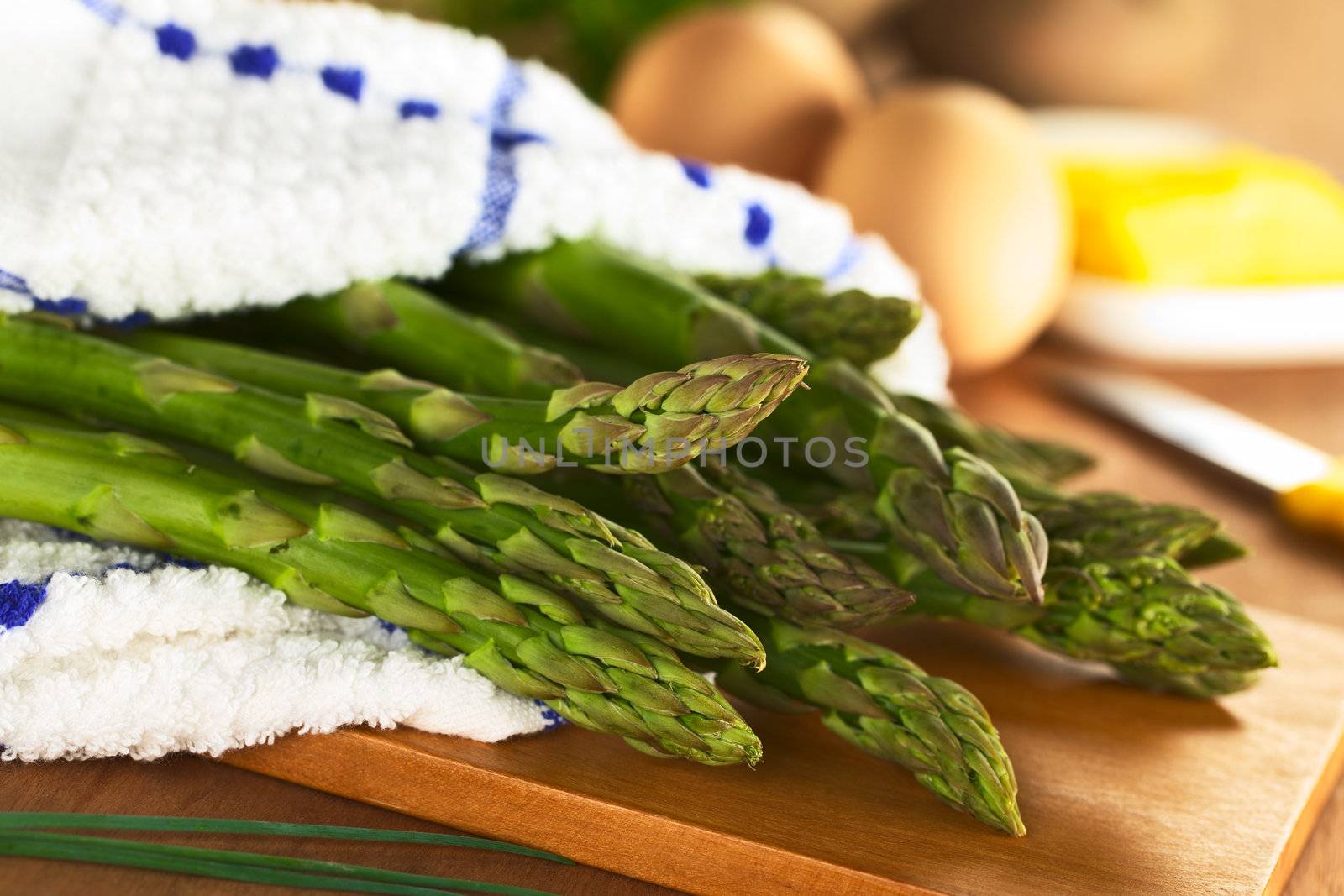 Raw green asparagus wrapped in a dish towel lying on a wooden board with eggs and butter in the back (Selective Focus, Focus on the three asparagus heads in the front)