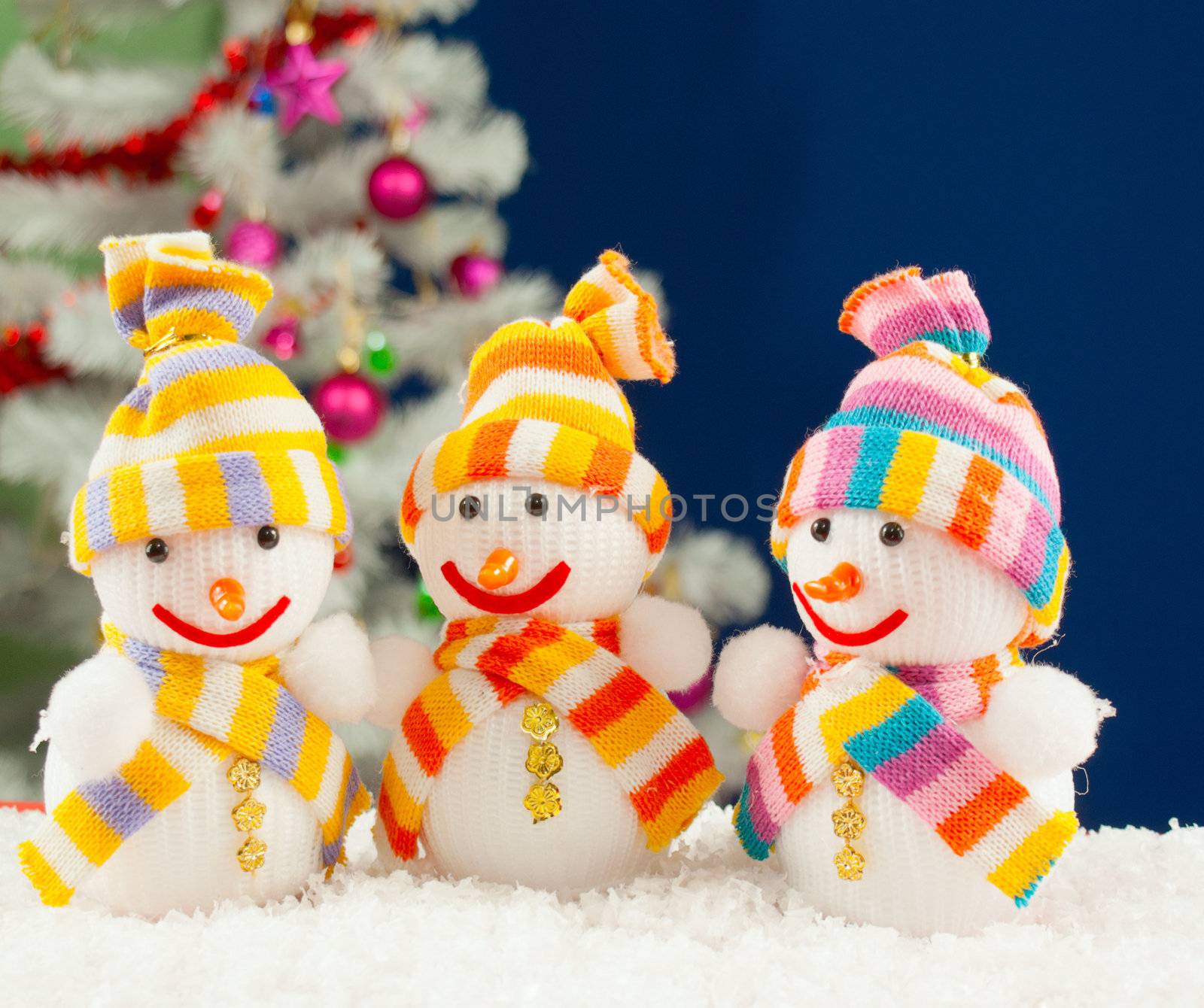 Three snowmen in front of the decorated white evergreen tree over the blue background