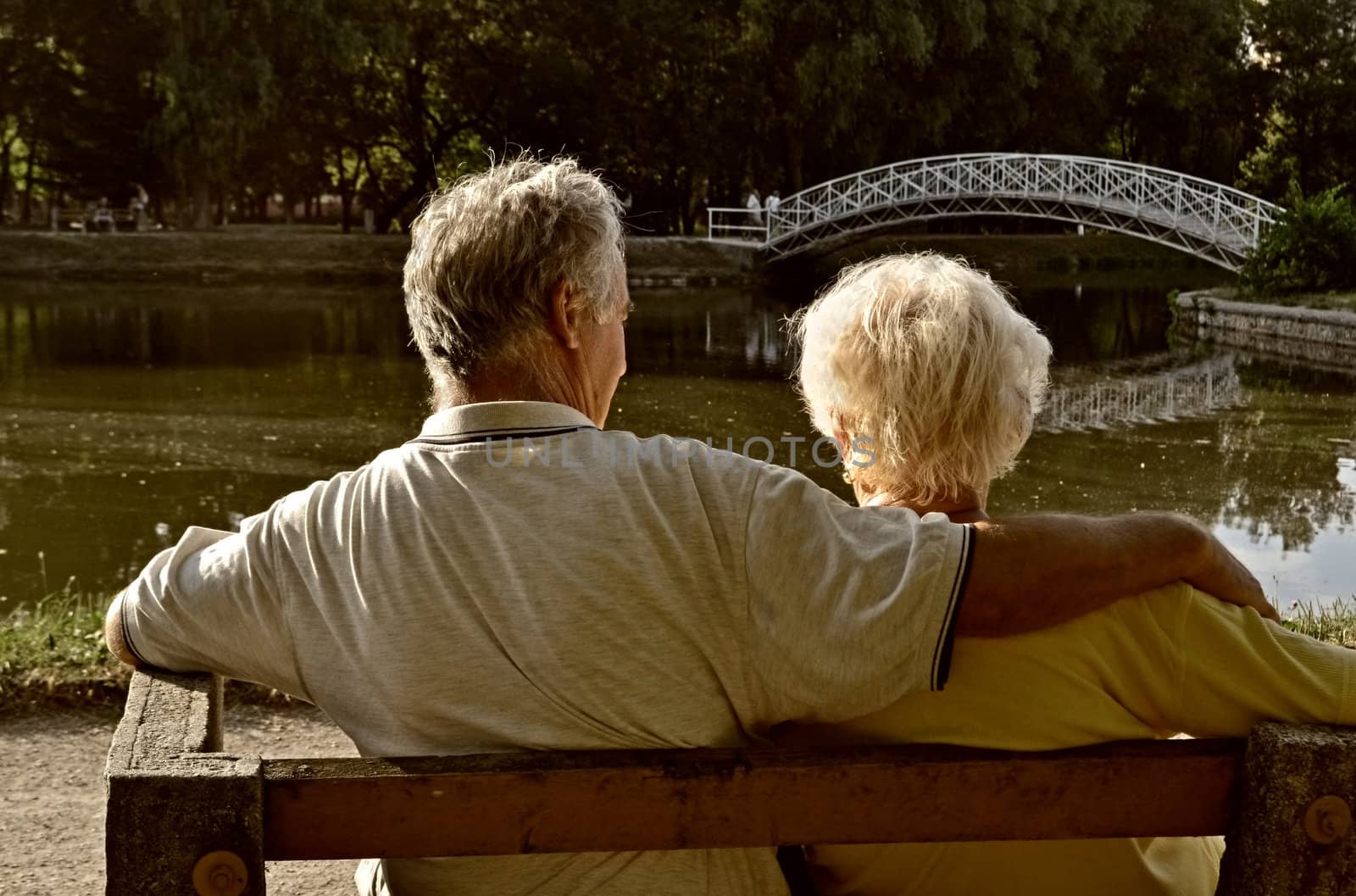 Senior couple relaxing, sitting on a pew by a small fishing lake in a leisure park, enjoying the afternoon sunlight.