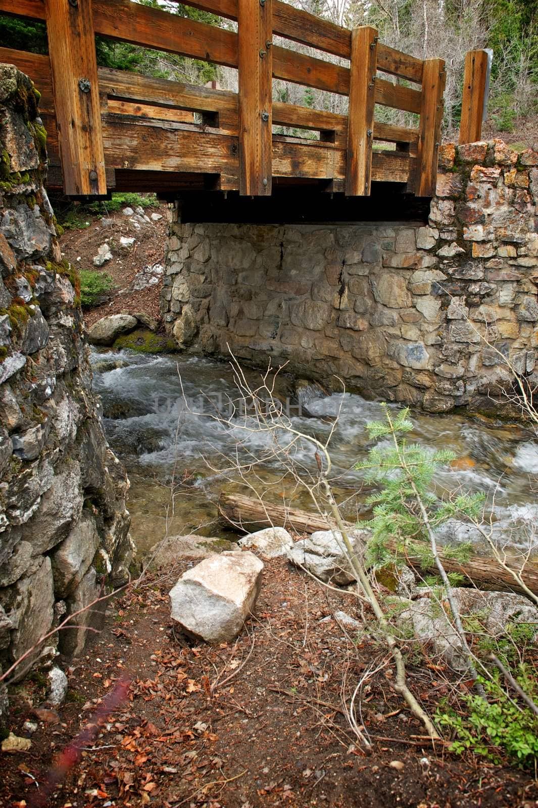 A weathered and worn wooden bridge stretches between two stone walls with a running stream