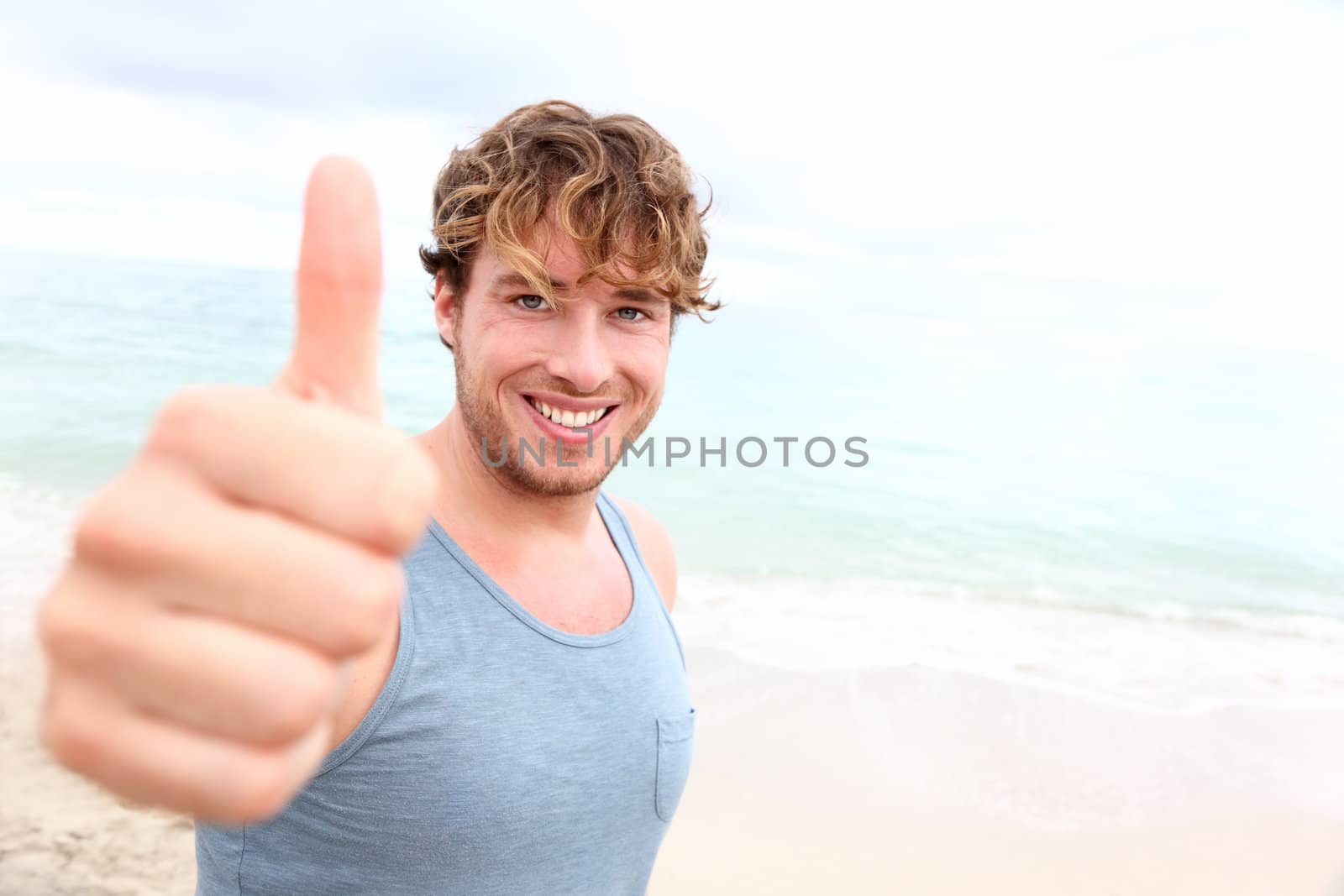 Young man thumbs up. Smiling happy sporty man giving thumbs up success sign to camera during training outside on beach. Handsome male fitness model in his 20s.