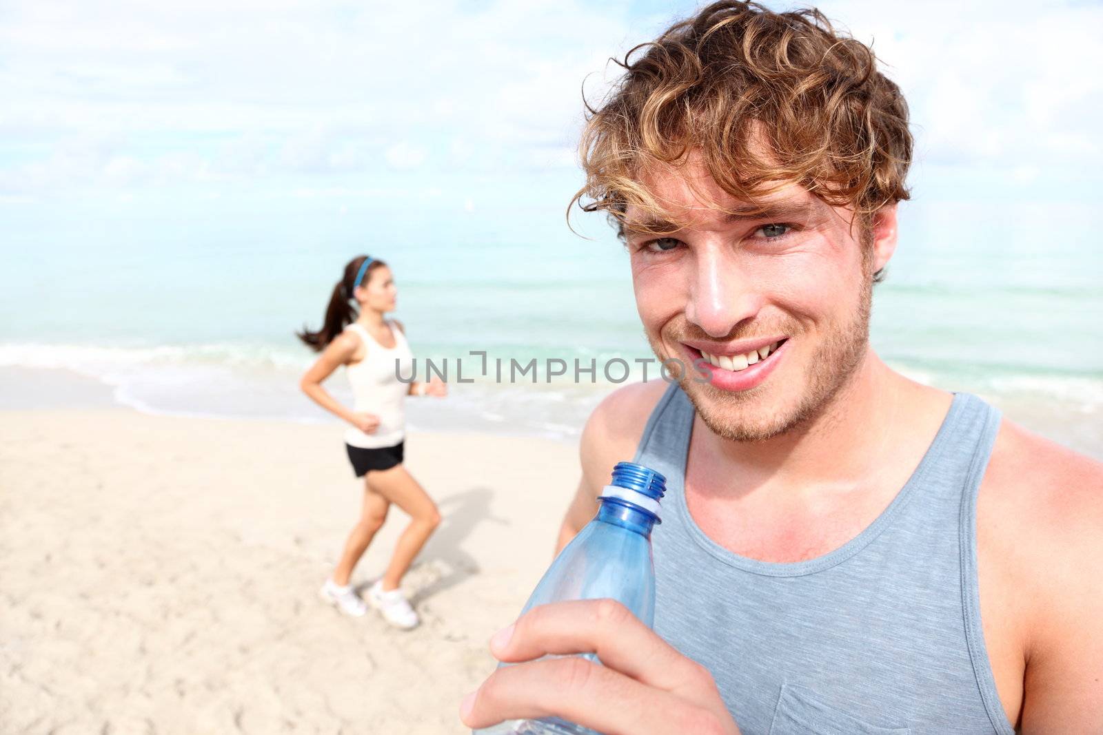 Running couple. Man drinking water smiling happy, woman runner in background. Young male fitness model in his 20s outdoors on beach