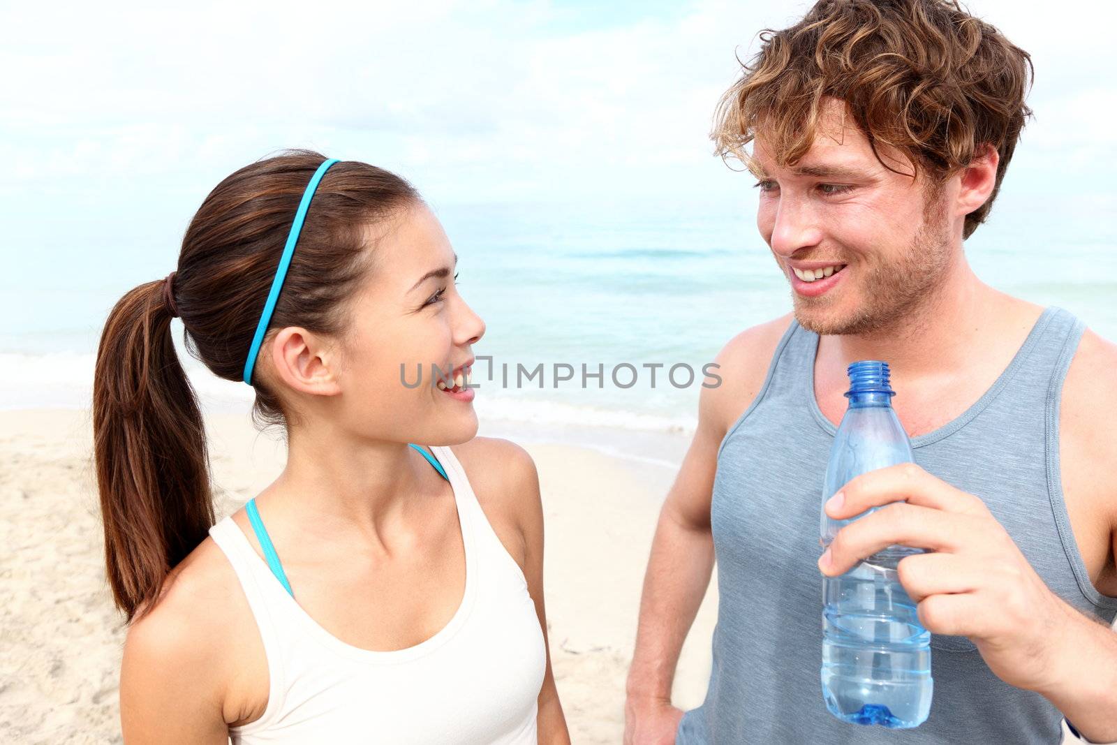 Fitness couple on beach talking relaxing and smiling happy after running training. Multiracial couple: Asian woman and Caucasian man drinking water