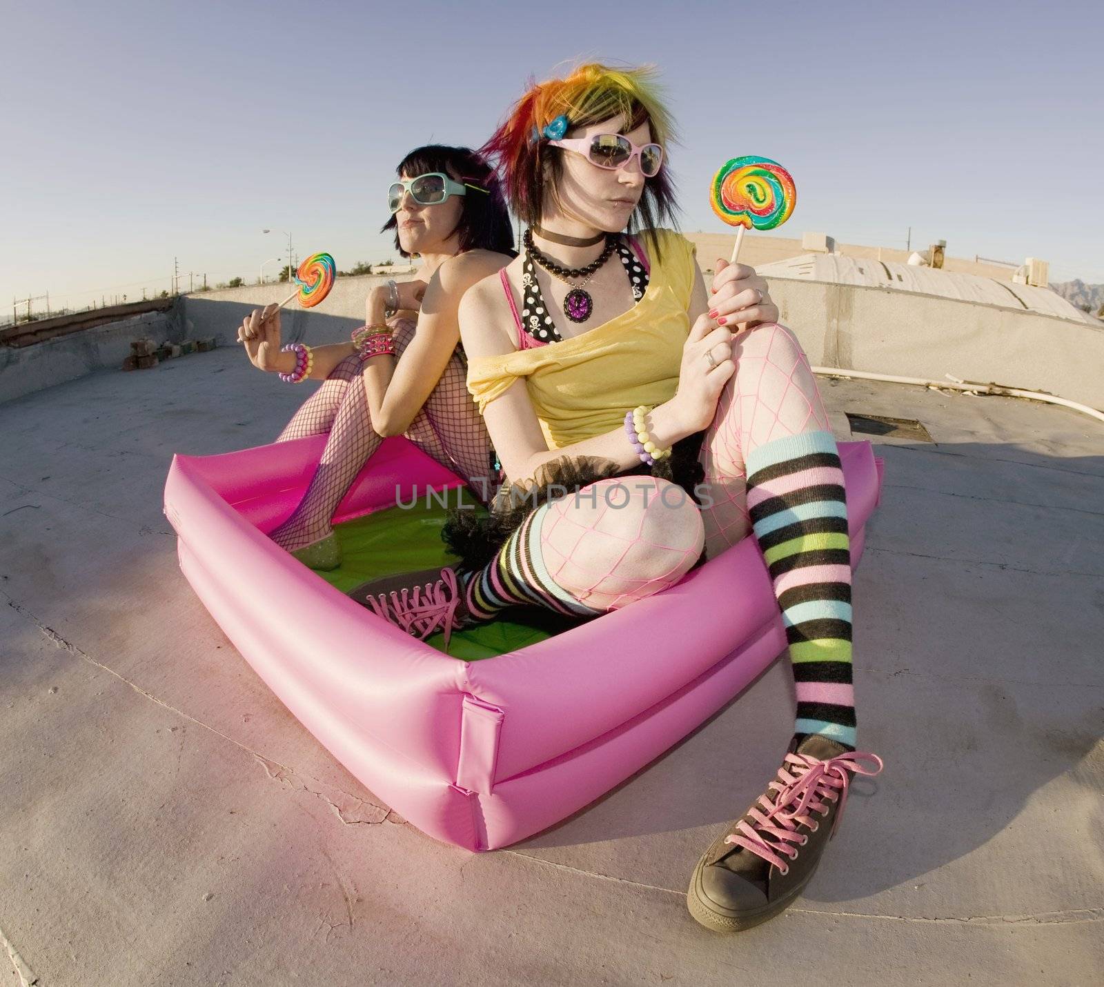 Fisheye shot of girls in brightly colored clothing in a plastic pool on a roof with sunglasses and lollipops
