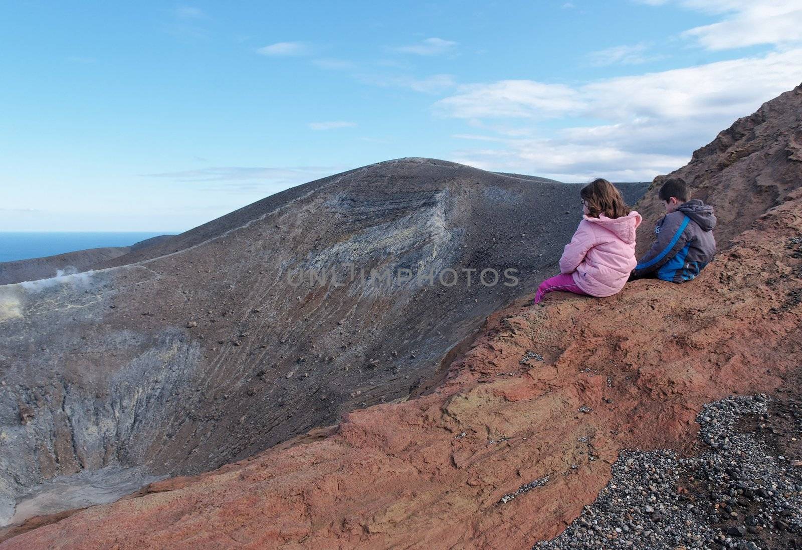 Girl and boy sitting on the rim of volcano crater of Vulcano island near Sicily, Italy