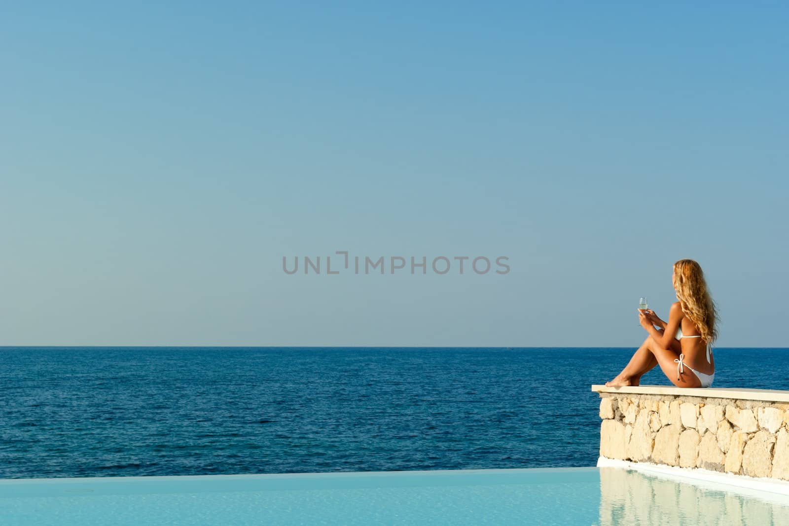 Beautiful woman in white bikini sitting near infinity pool and looking to the sea