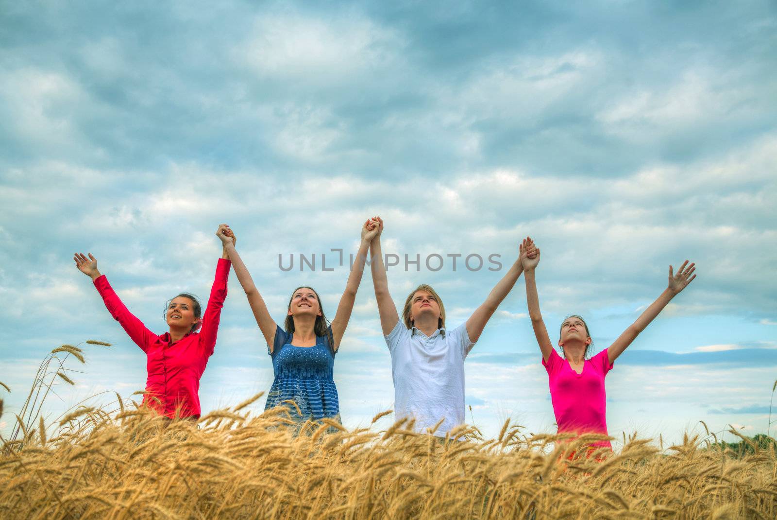 Four young people staying with raised hands at a wheat field at sunset time