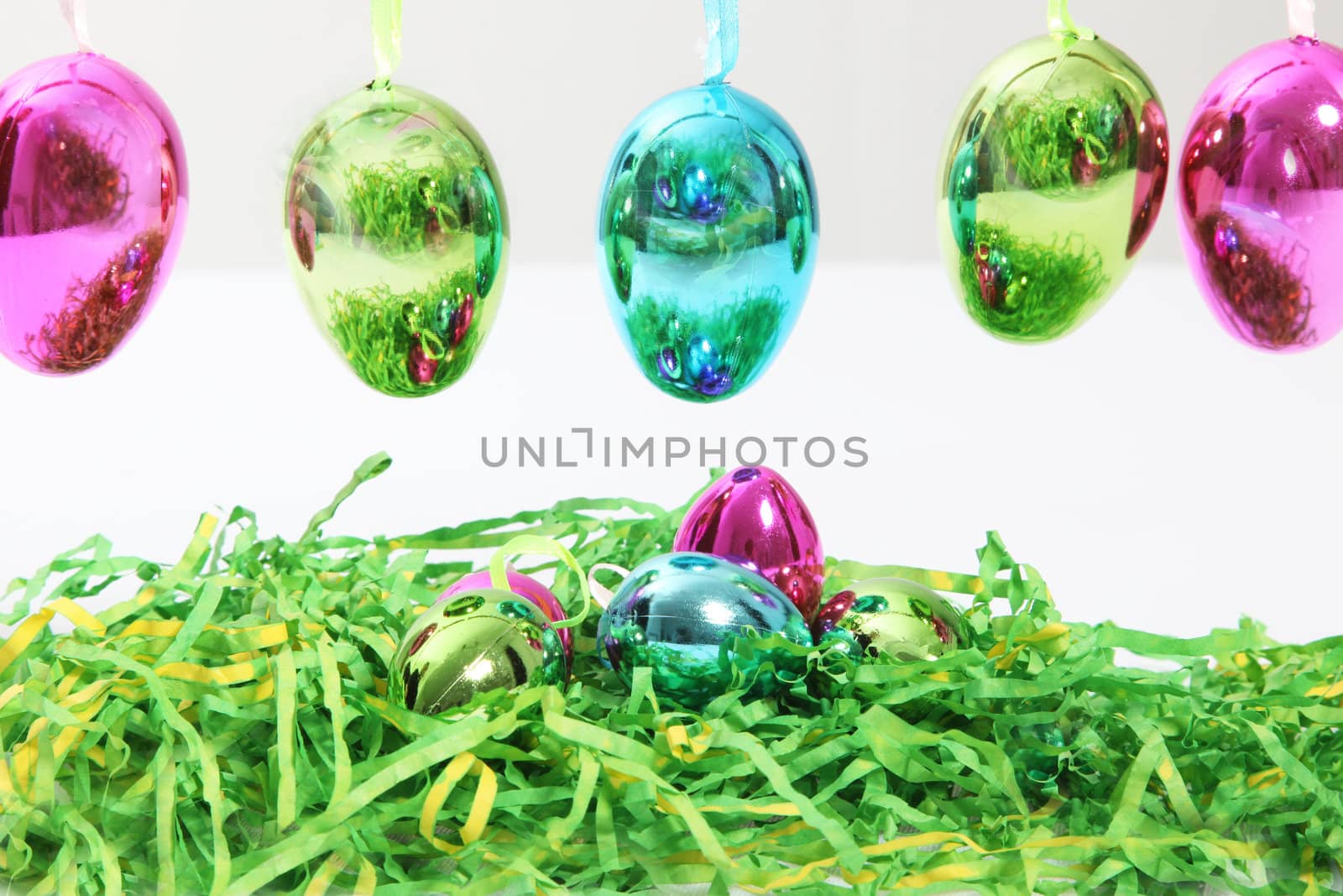 Colourful shiny metallic Easter Eggs hanging in a row above additional eggs nestling in a bed of green straw for a vibrant seasonal greeting card