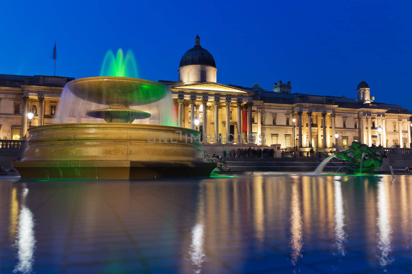 Trafalgar Square in the evening, the National Gallery. In the foreground - illuminated fountain and pool, reflecting the sky and lights. London, UK