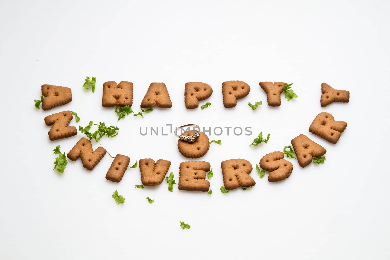 Happy anniversary wording by brown biscuits, ring and green leaves on white surface