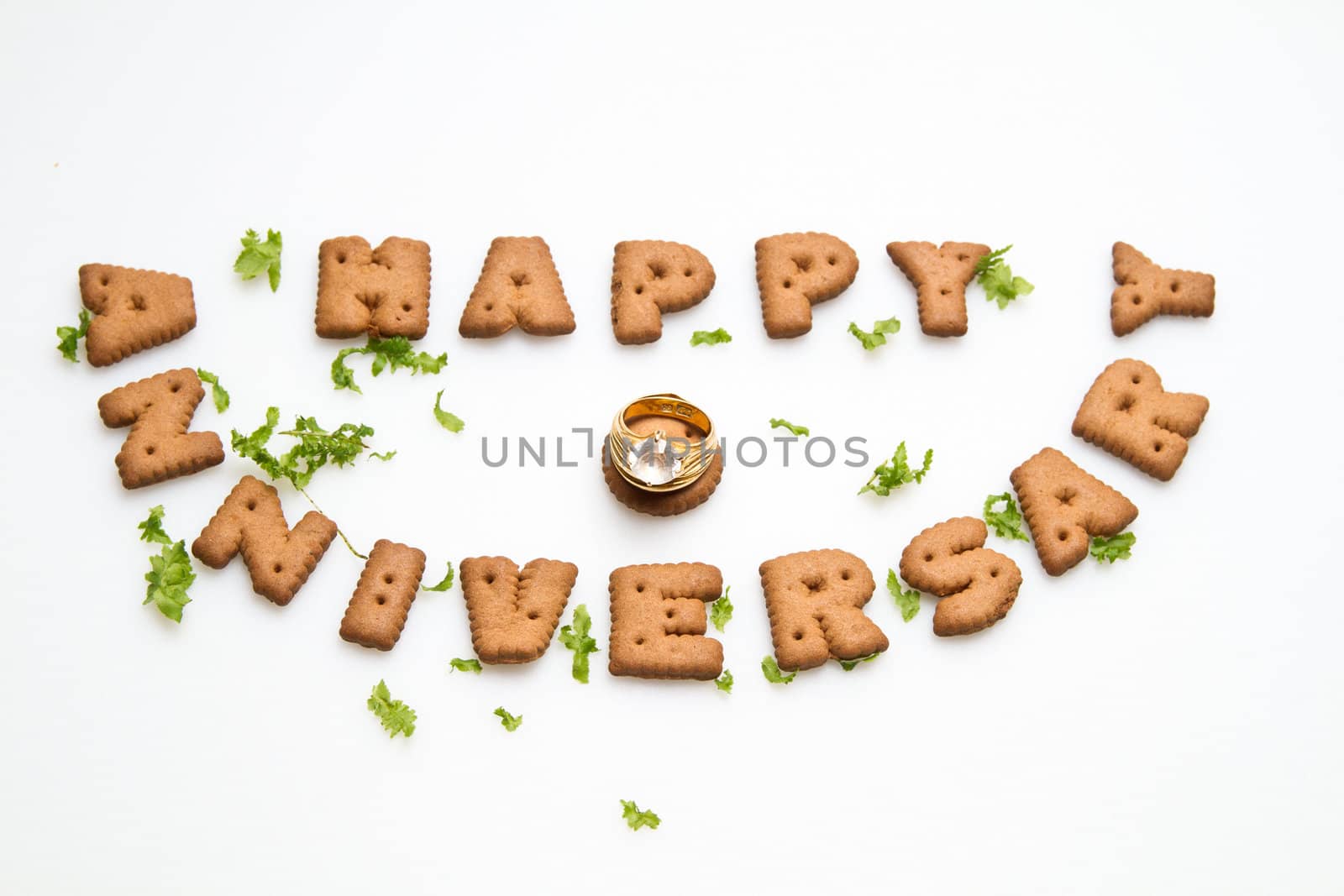 Happy anniversary wording by brown biscuits, diamond ring and green leaves on white surface