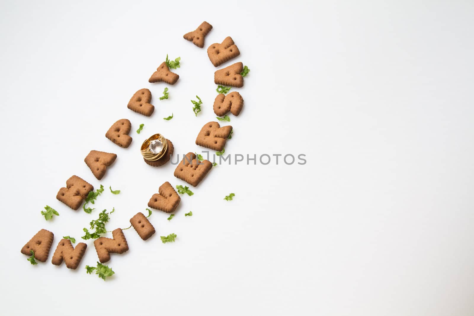 Happy anniversary wording by brown biscuits, diamond ring and green leaves on white surface slanting position