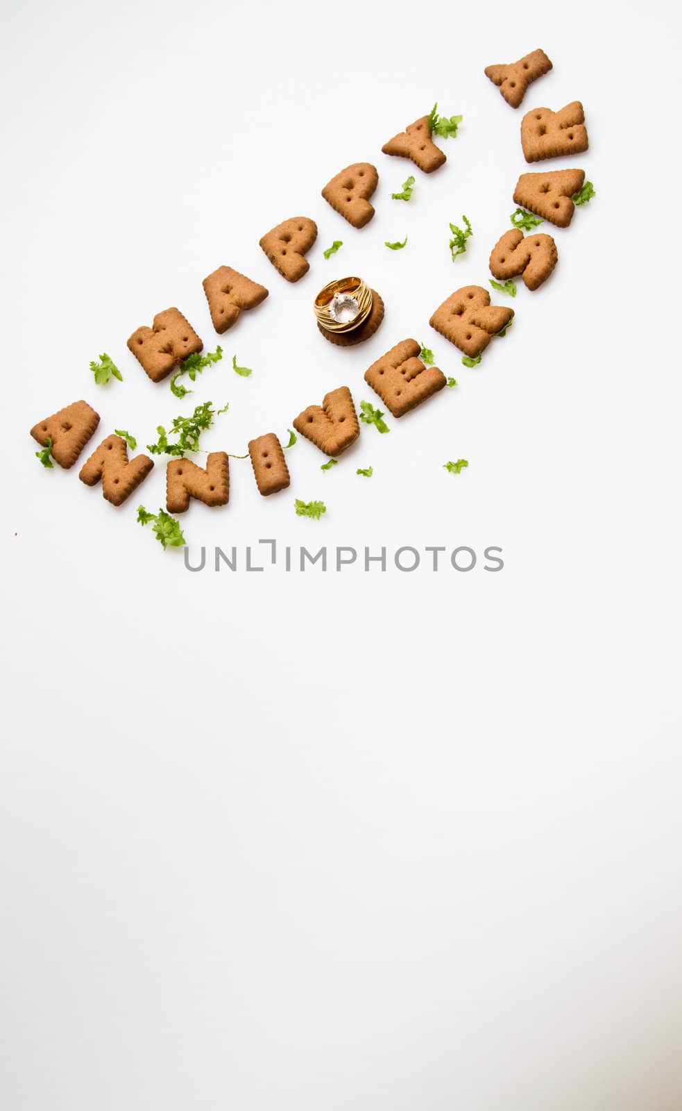 Happy anniversary wording by brown biscuits, diamond ring and green leaves on white surface slanting position