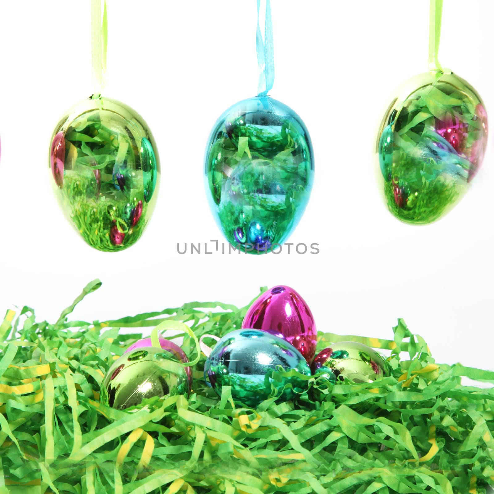 Row of colourful shiny Easter Eggs hanging on ribbons above additional eggs nestling in green straw isolated on white colourful eggs on white background