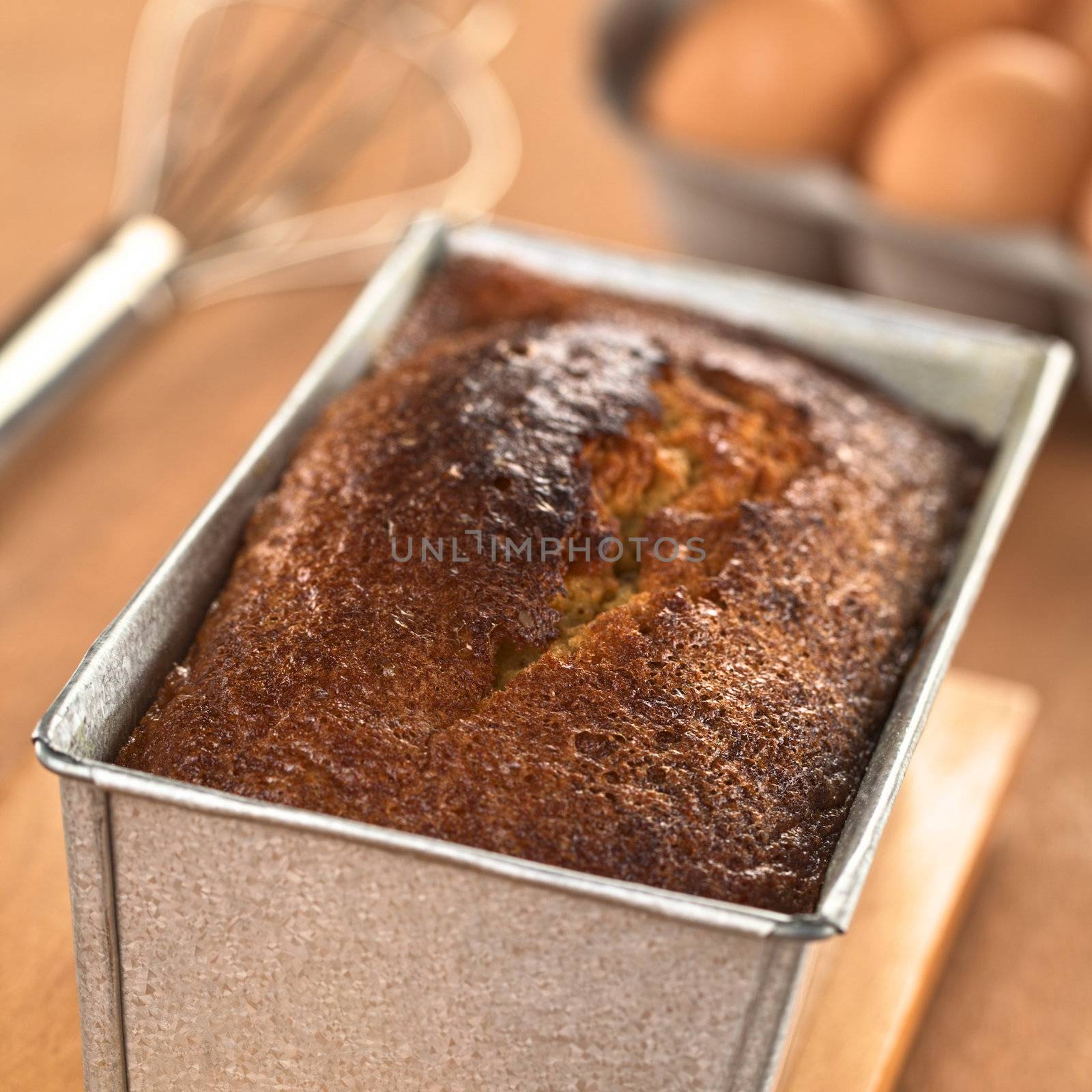 Freshly baked pound cake in baking pan with eggs and beater in the back (Selective Focus, Focus one third onto the surface of the cake) 