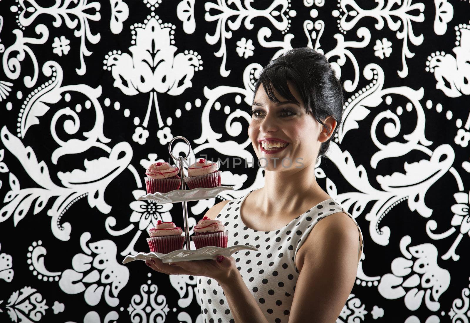 young woman is stylist 60's inspired clothing, with a big smile, holding a plate of velvet cupcake