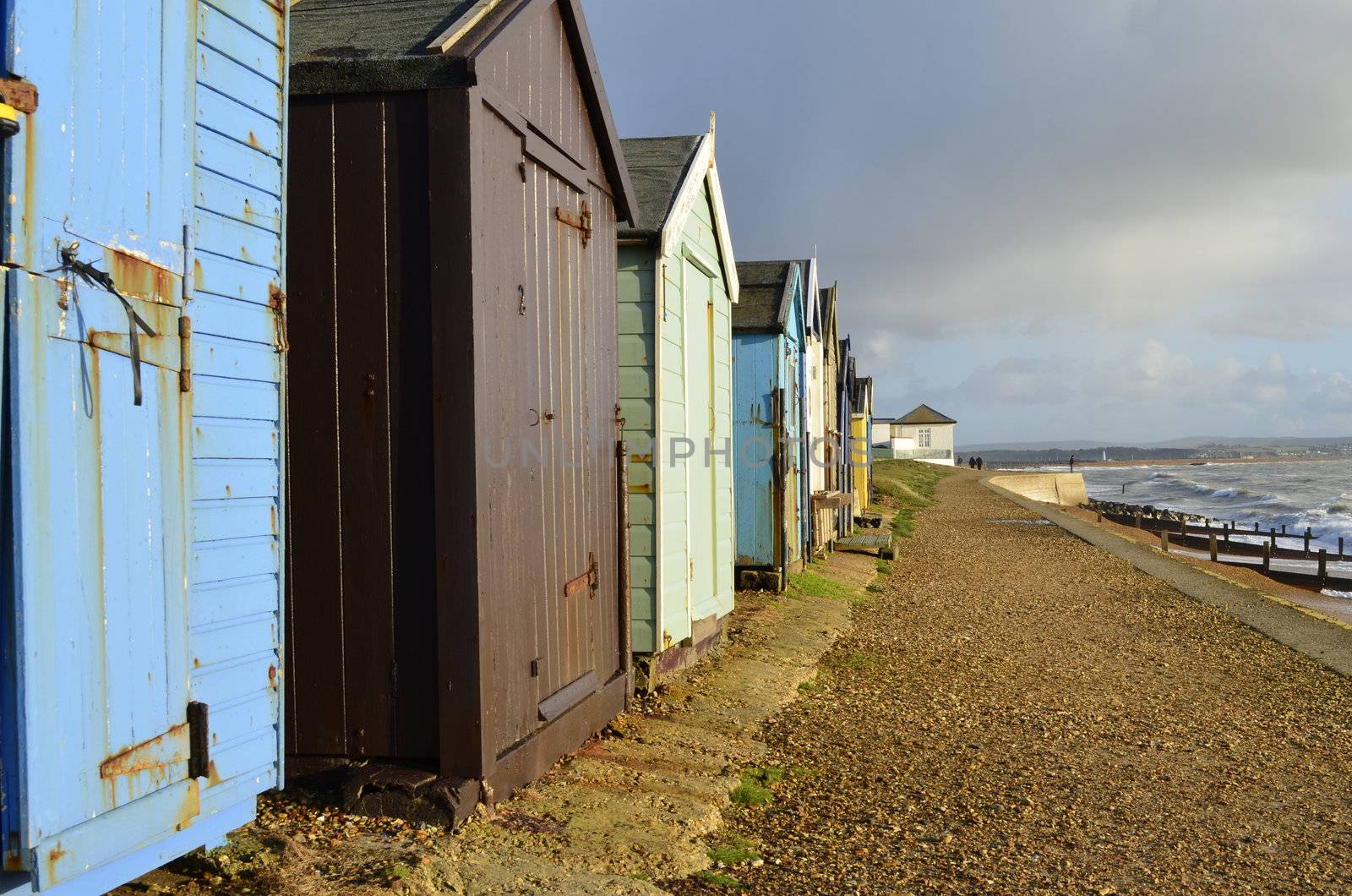 Old coastal beach huts looking our to the wintery sea in full colour with couple walking in distance.