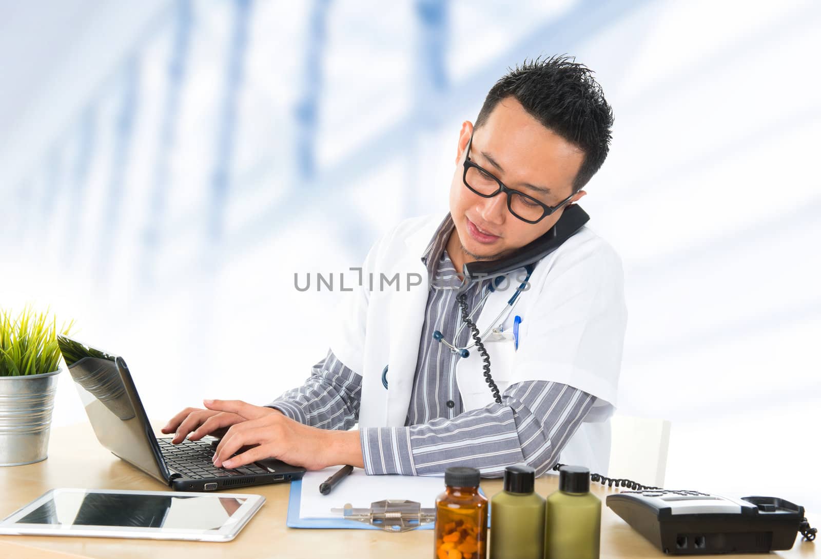 Busy Southeast Asian medical doctor on the phone and typing on notebook, sitting in front working desk in hospital.