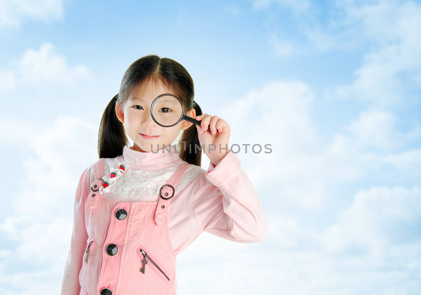 A little Asian girl peers at the camera through a magnifying glass in summer day, blue sky background