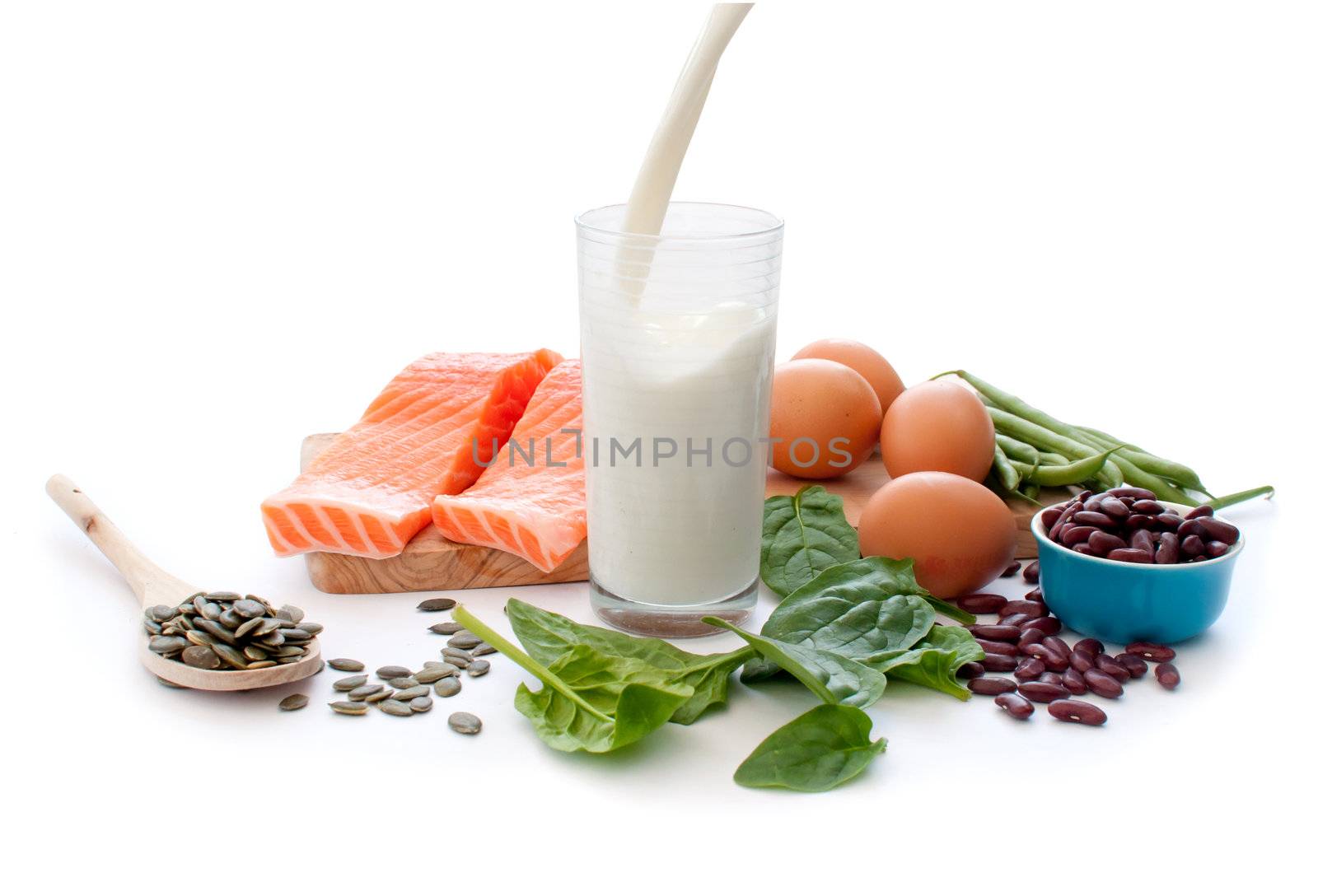 Protein rich foods including eggs, spinach leaves and pumpkin seeds with a glass of pouring milk in the middle
