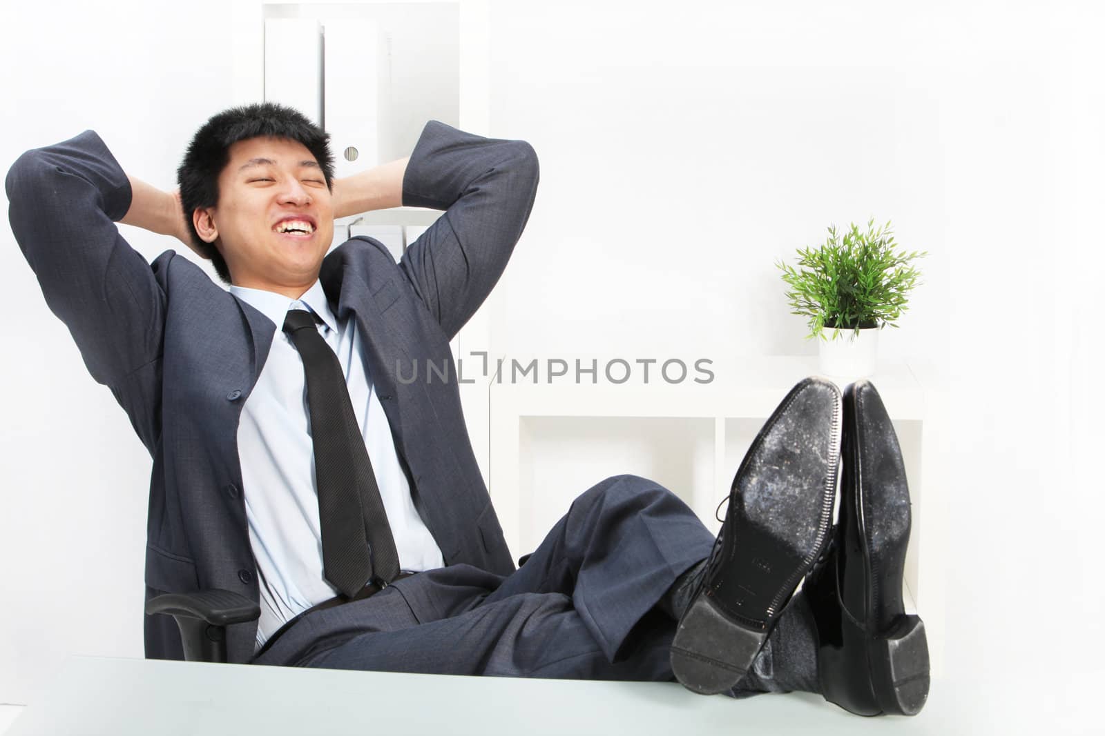 Asian businessman relaxing with his feet up on top of his desk and his hands clasped behind his head smiling happily to himself