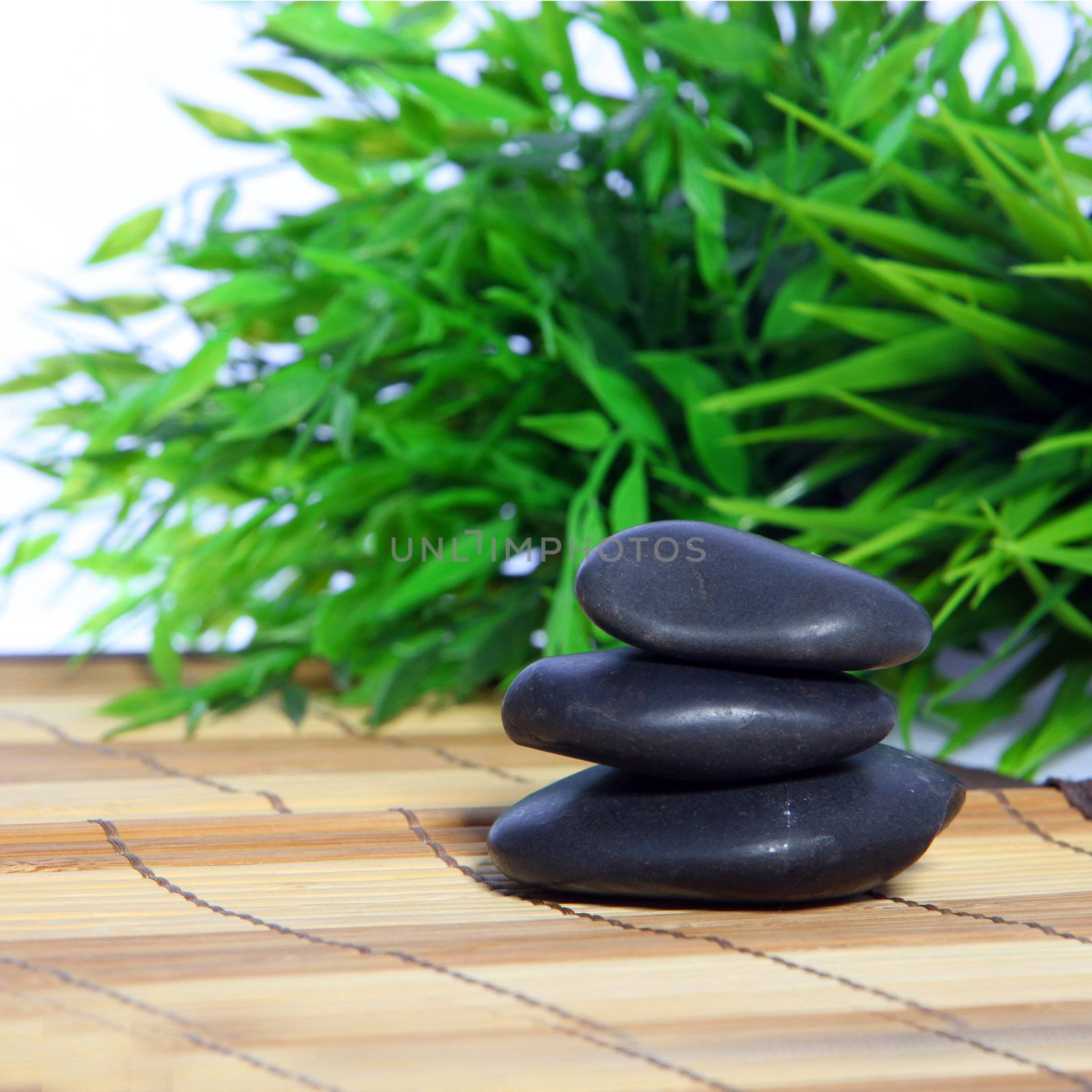 Stack of smooth black basalt massage stones on a reed mat in a spa with a backdrop of bamboo leaves