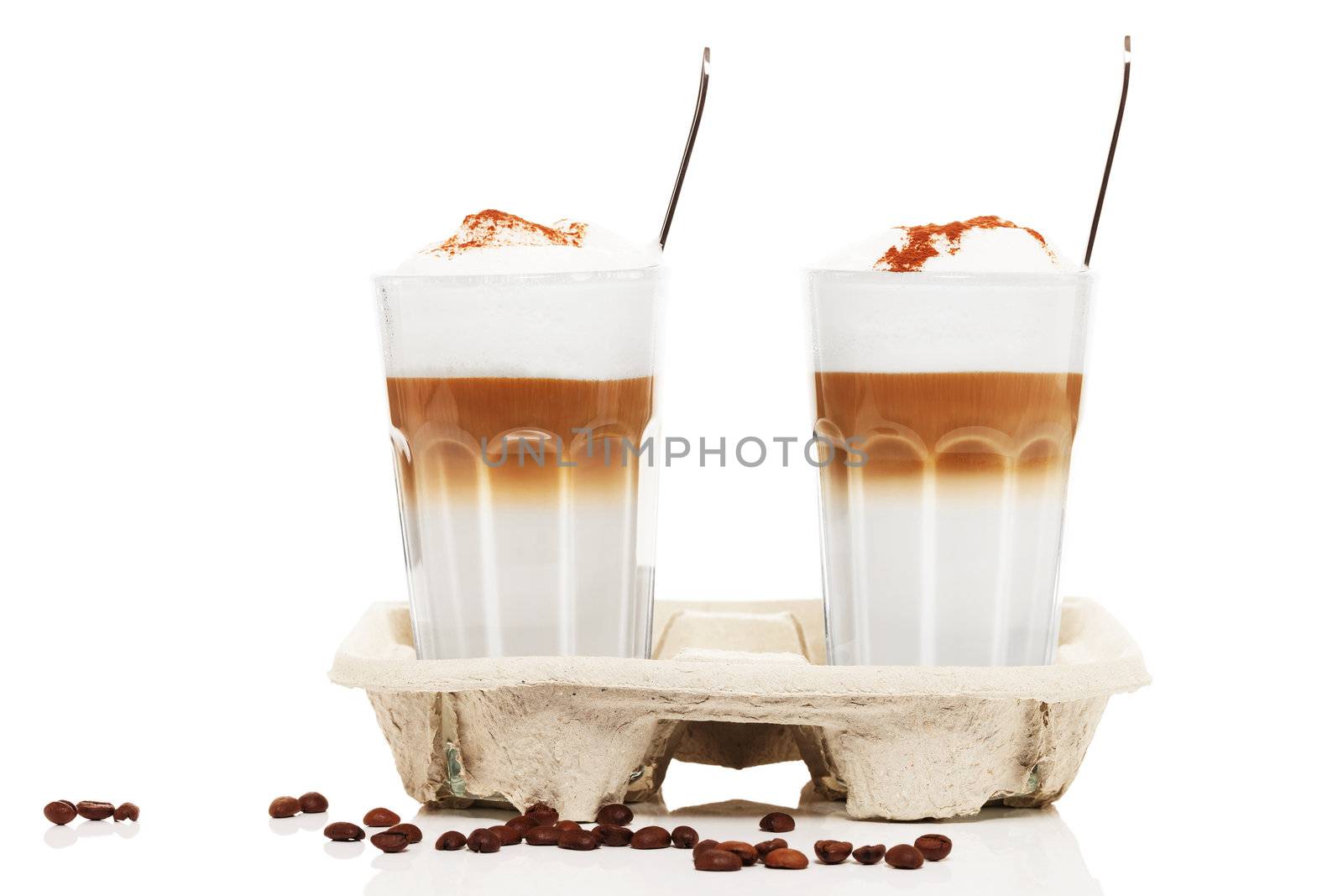 two latte macchiato with chocolate powder in a cupholder with coffee beans  and spoons on white background