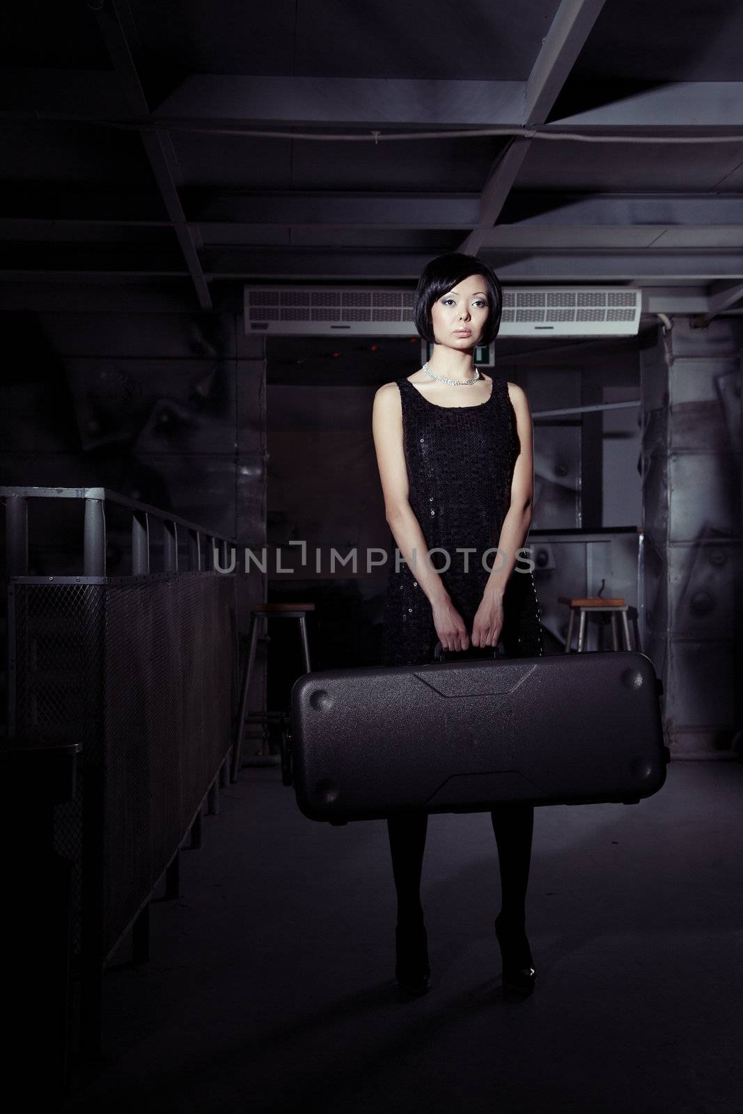 Small brunette lady holding big suitcase in the dark airport terminal