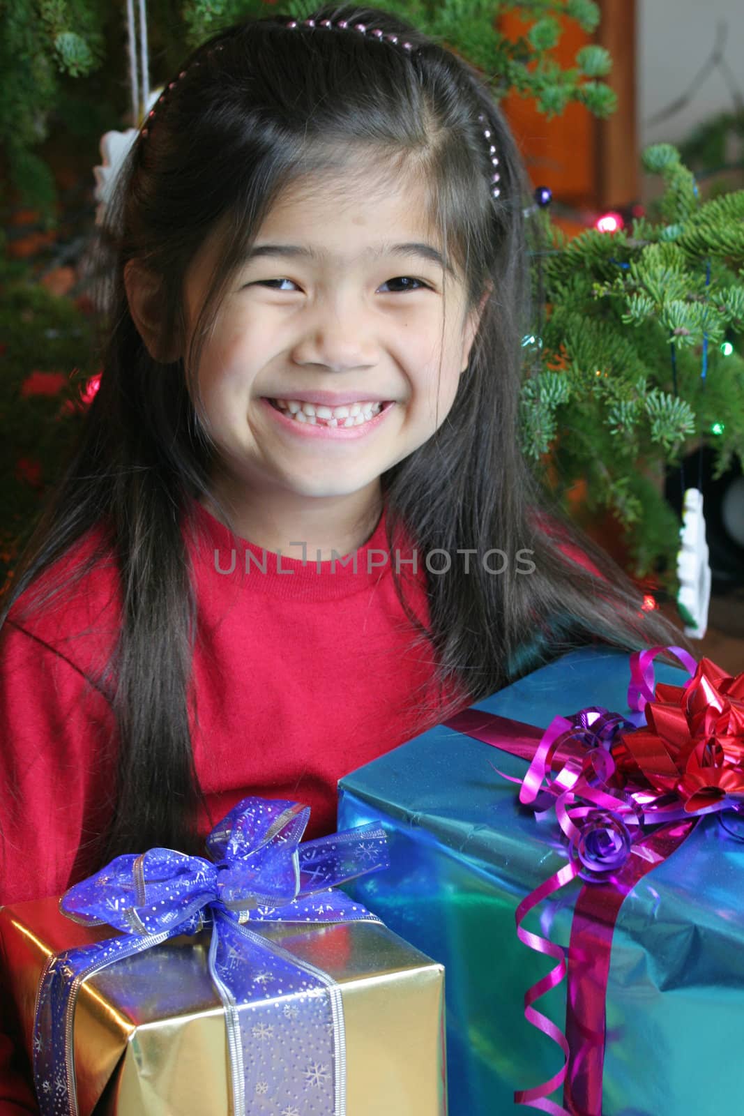 Happy little girl holding her Christmas presents by the Christmas tree. Part Asian, Scandinavian heritage.