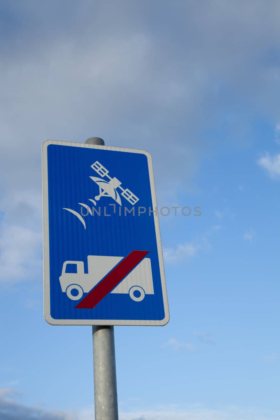 A sign with a lorry, truck, and red slash with a satellite tracking symbol above, on a blue background against a blue sky with cloud