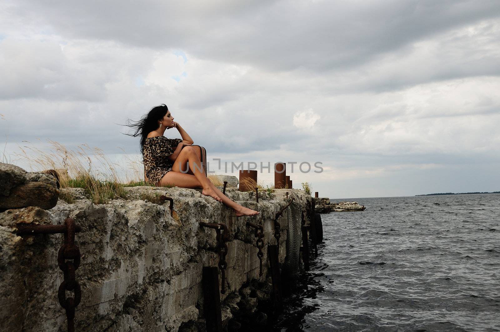 A beautiful brunette woman in mini dress and bare feet sits alone on a windswept stone wall bordering the sea