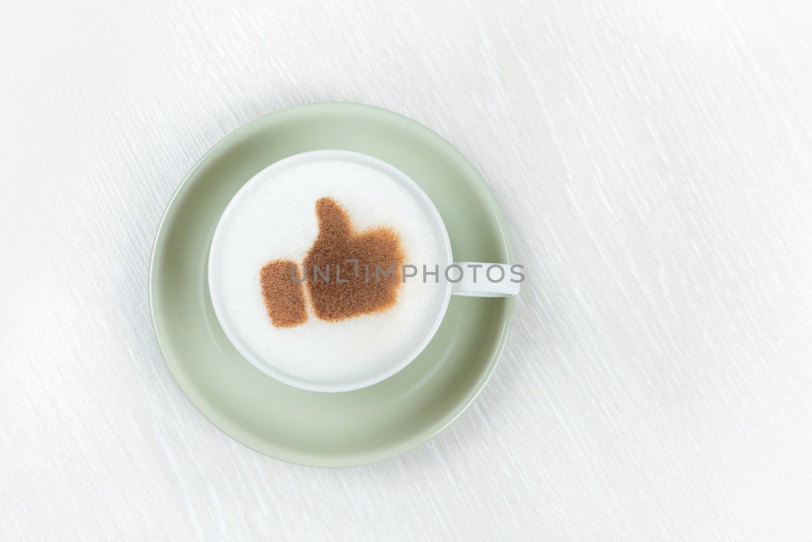 Top view of a cup filled with frothy cappuccino with a brown thumbs up sign in cocoa powder depicting success, praise and approval on a white table top with copyspace.