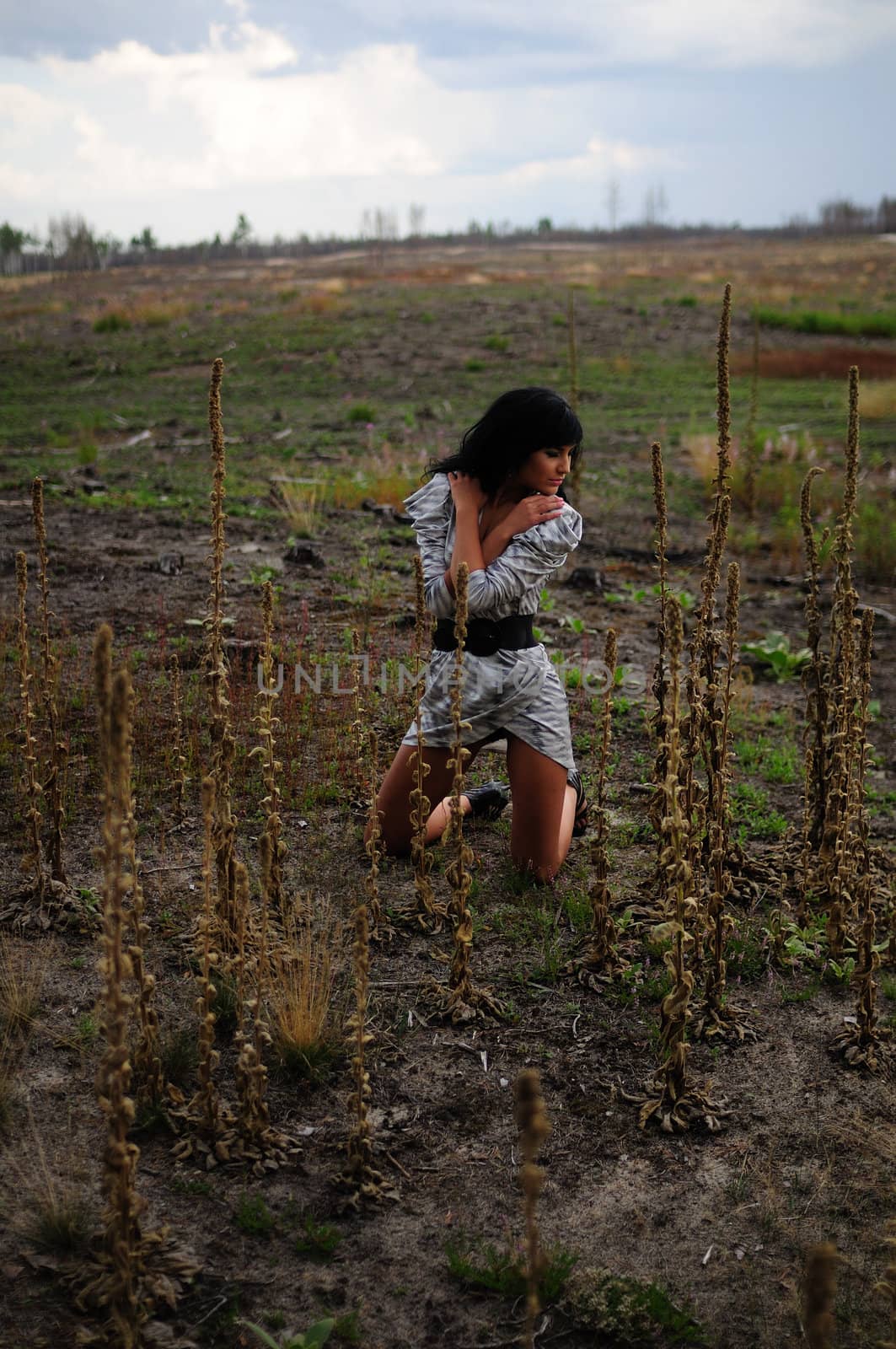 Beautiful woman in a dress kneeling in a vast open field with her hands crossed over her chest.