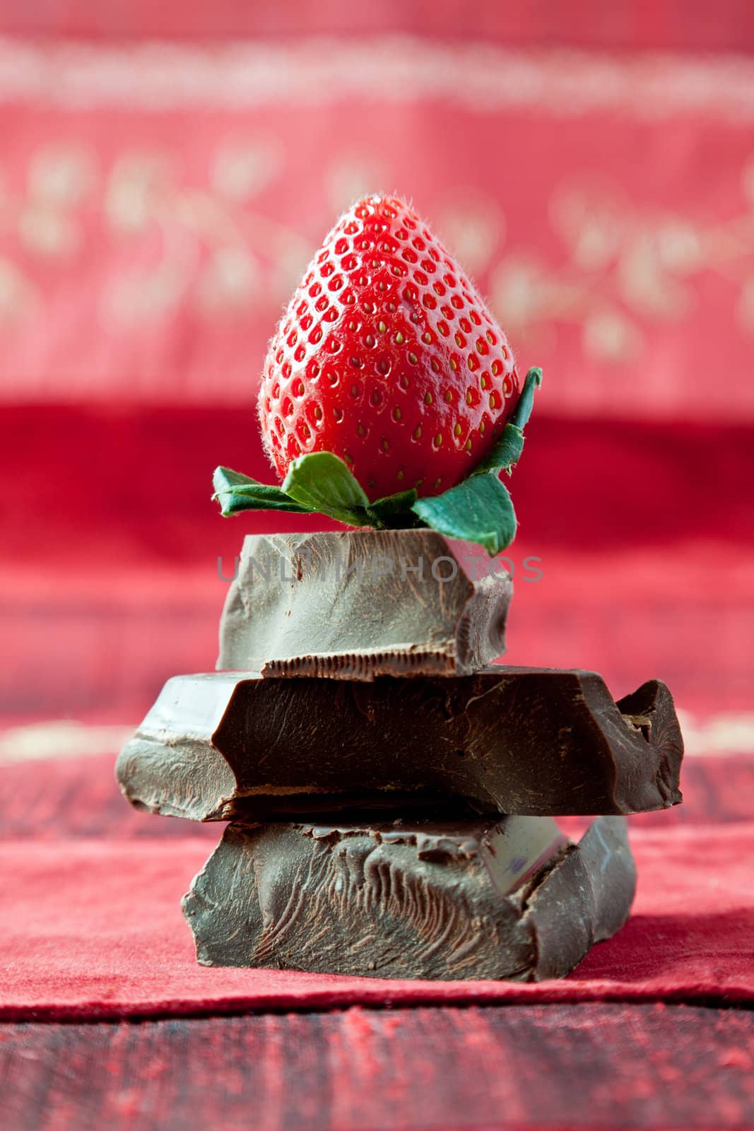 Strawberry on top of pile of gourmet thick dark chocolate bar chunks. Red textured background.