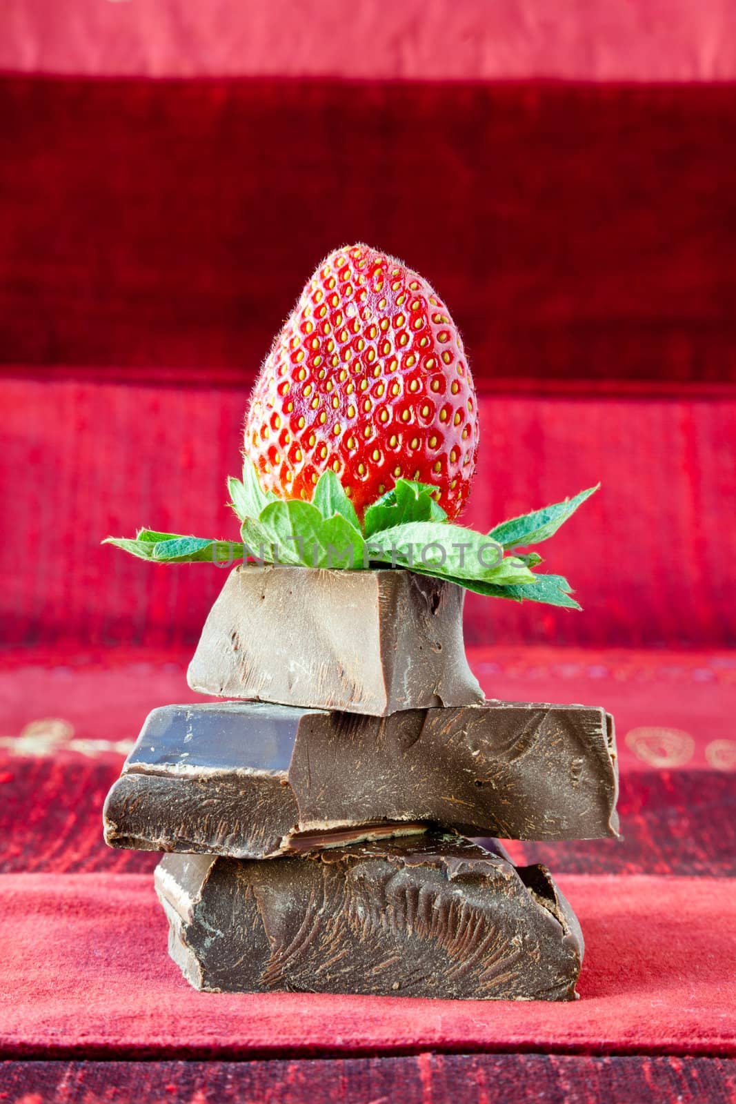Strawberry on top of pile of gourmet thick dark chocolate bar chunks. Red textured background.