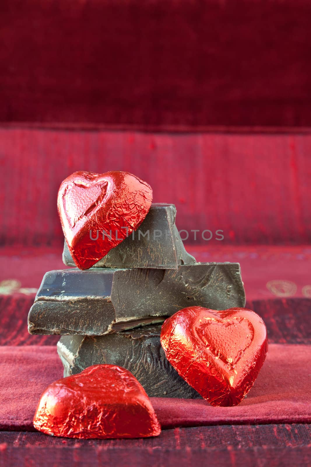 Red foil wrapped candy hearts arranged on top of pile of gourmet thick dark chocolate bar chunks. Red textured background.