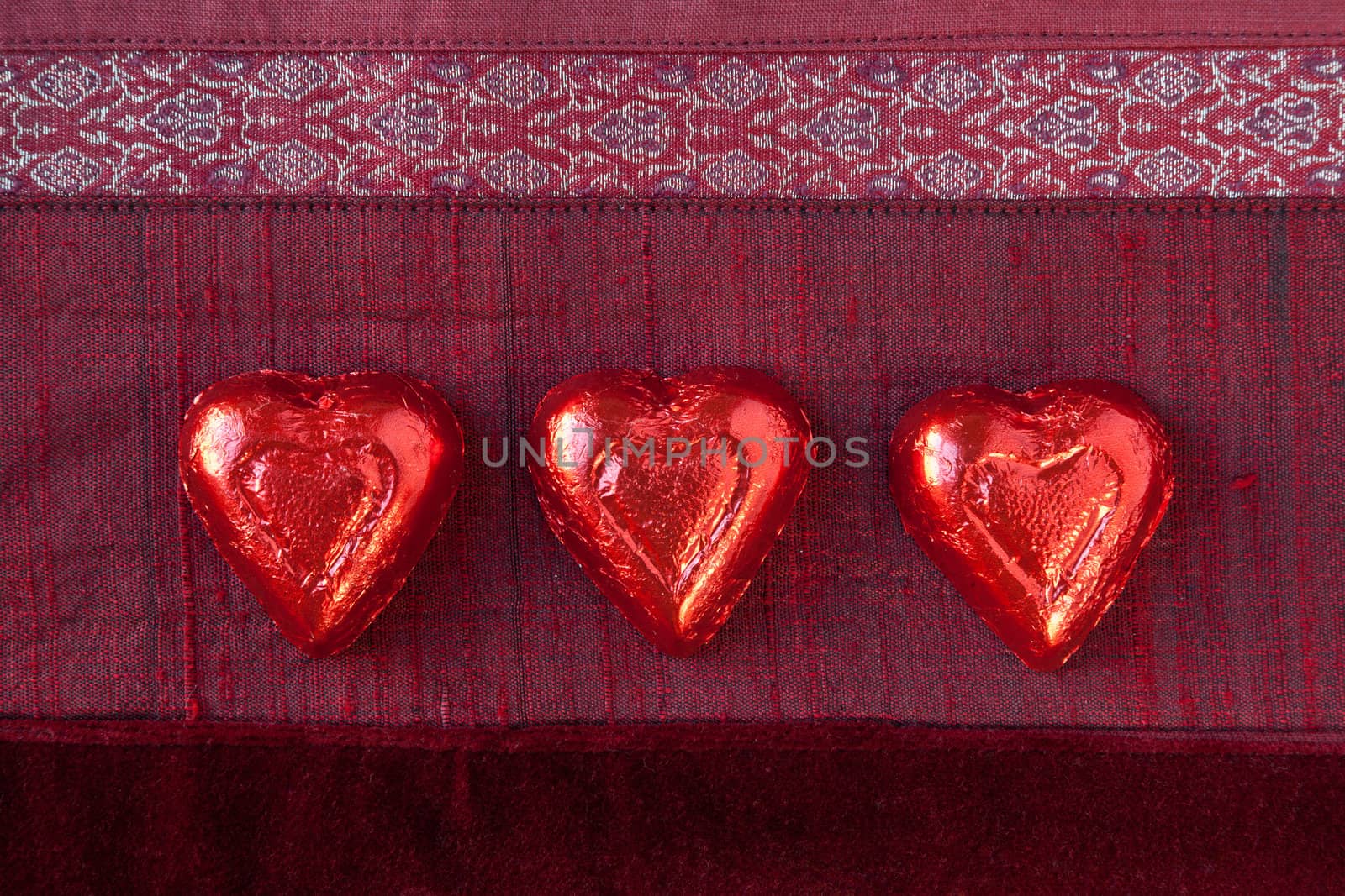 Three red foil wrapped candy hearts in a row on red textured background.