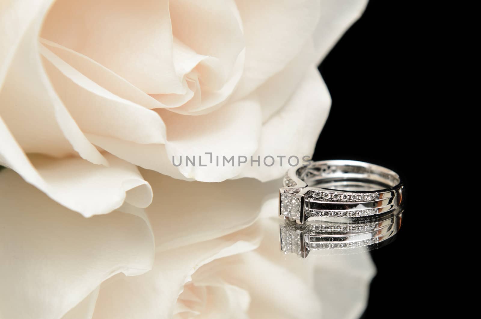 A diamond engagement ring with a white rose, shot with a reflection on a black background.