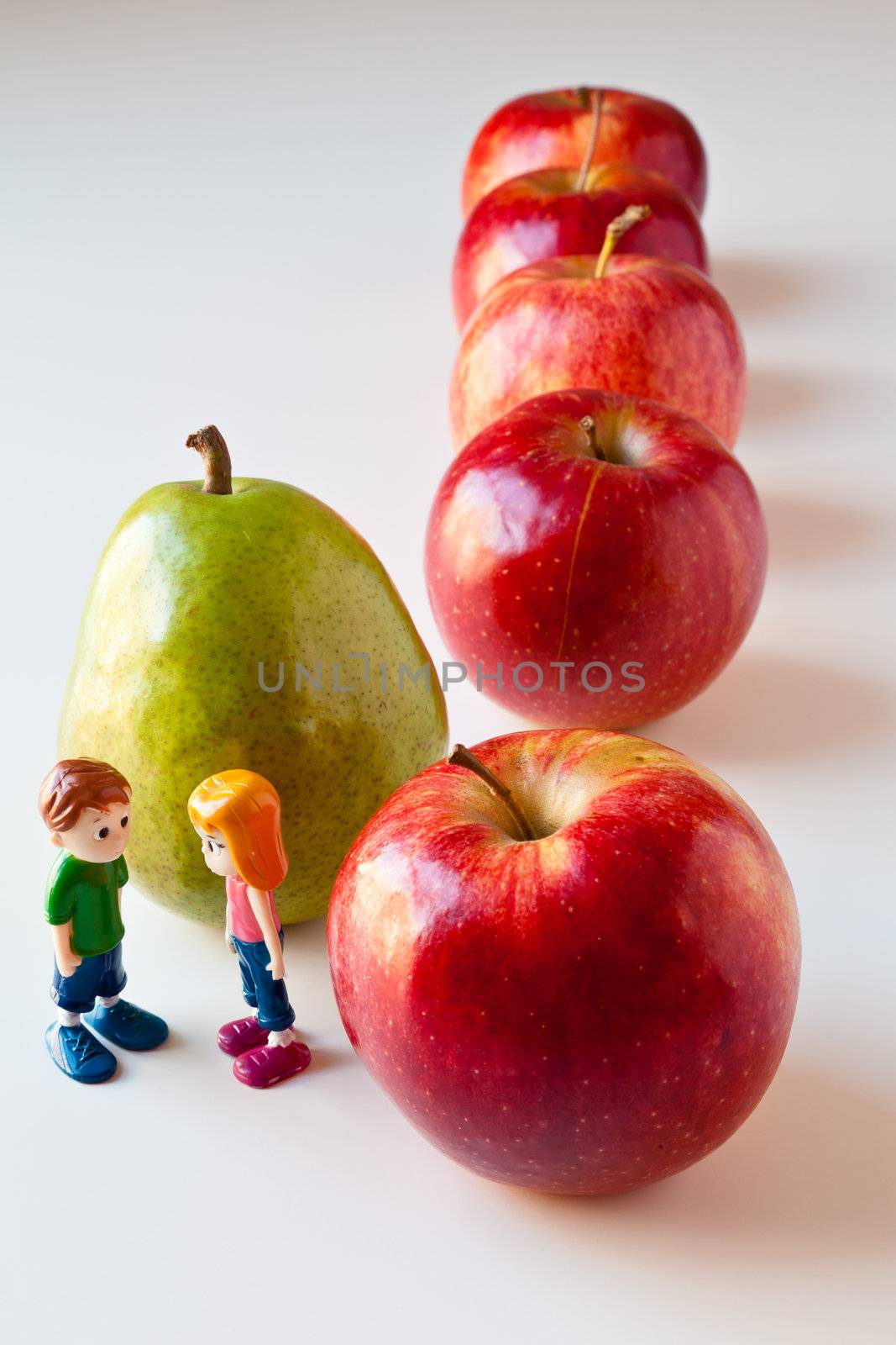 Toy girl and boy discuss nutrition and healthy choices in front of green pear standing out from a line of red apples. The concepts depicted in this image are nutrition, good food choices, balanced diet, good for you, being different, unique, stick out, being singled out.