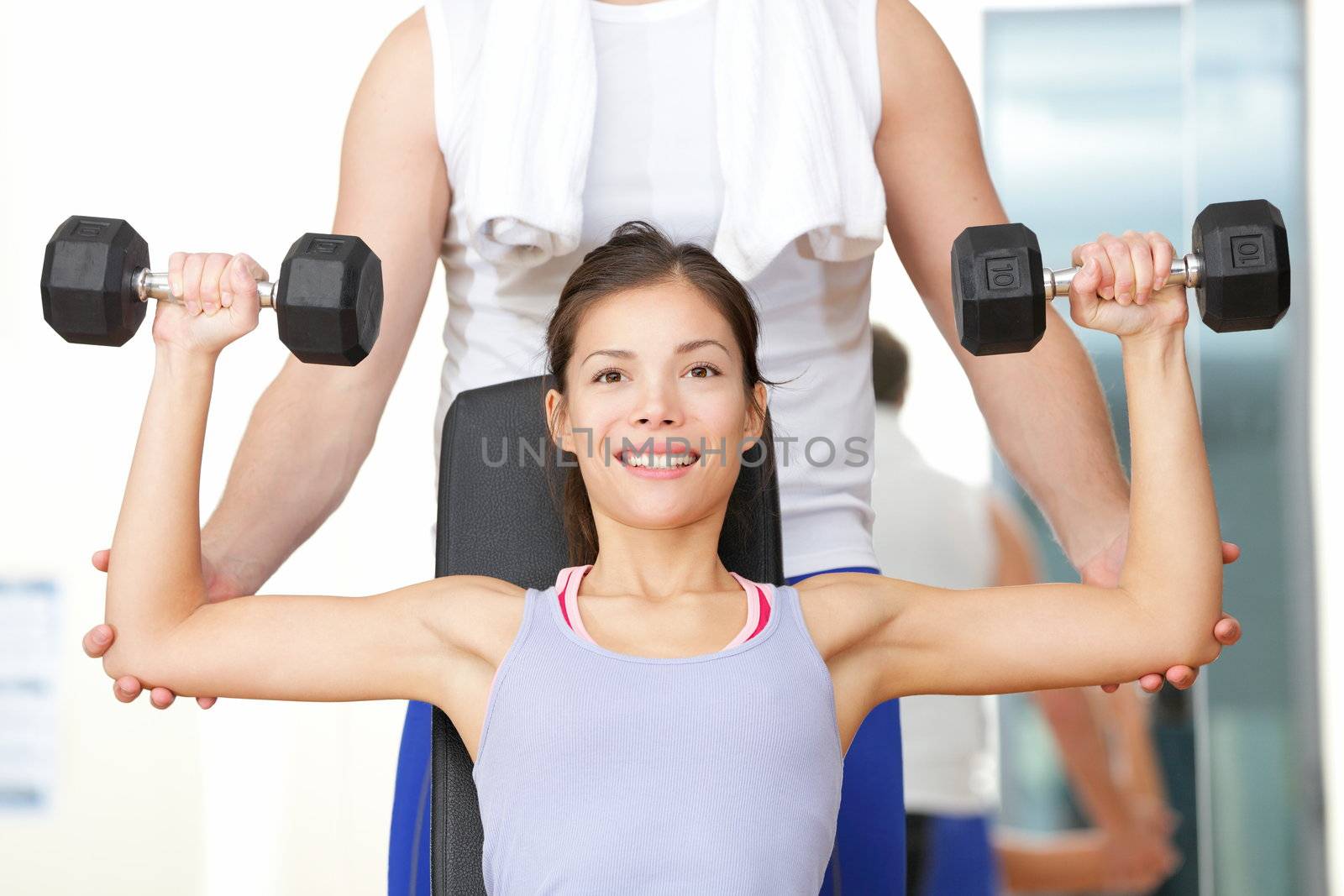 Gym fitness people - woman lifting weights with help from instructor and fitness trainer in gym. Beautiful smiling happy fit female fitness model training shoulders.