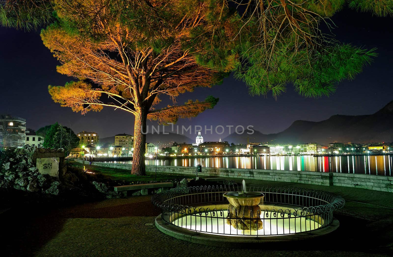 Lakeside city at nigh with illuminated fountain in foreground - Como Italy