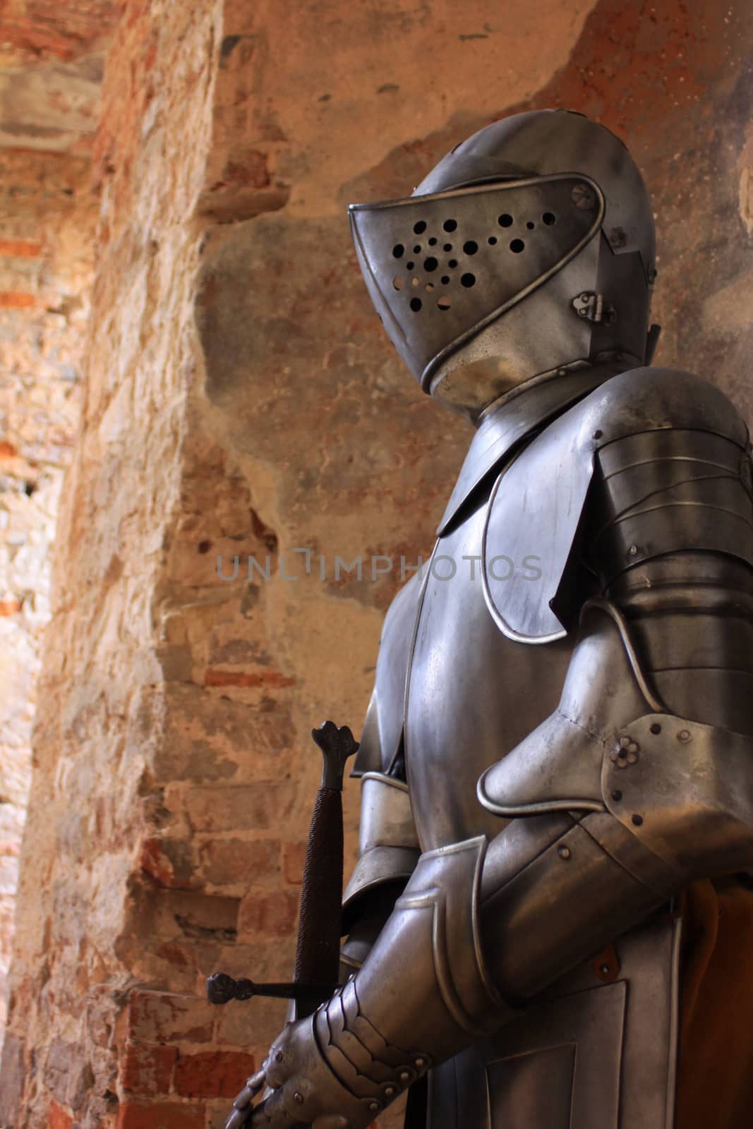 A medieval suit of armour with sword set against a stone bricked wall background.