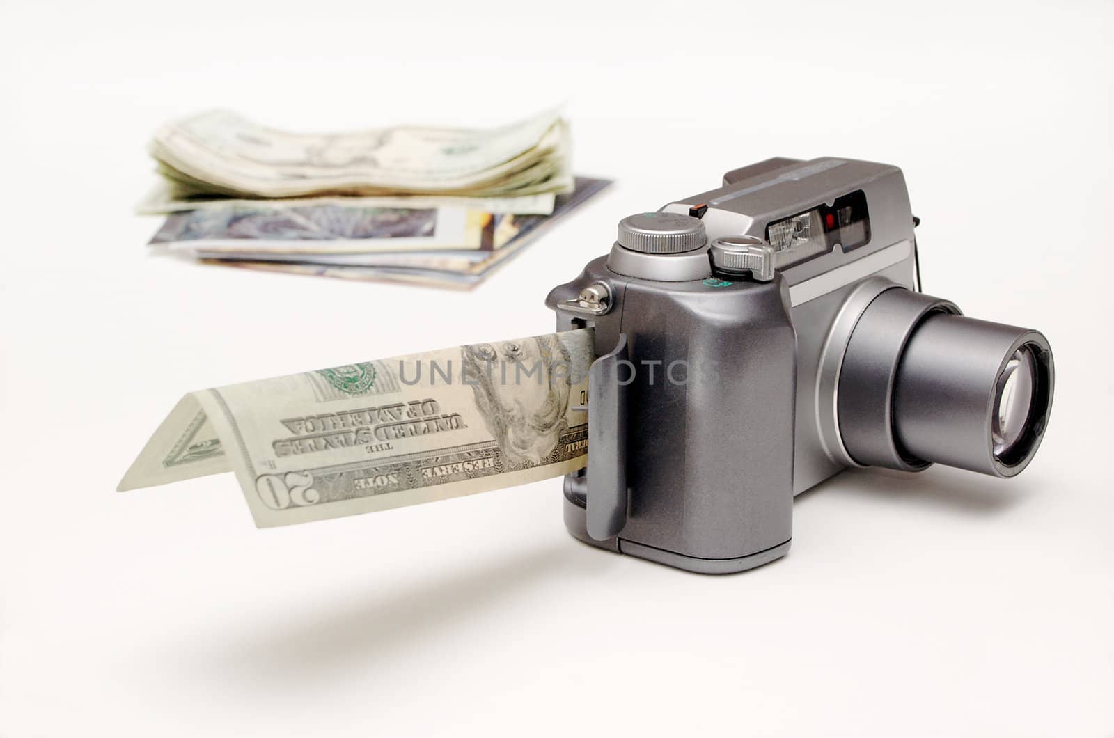 A digital camera, with the memory card slot open and a  bill sticking out of it, sits in front of a small pile of photos and money (which are defocused to show depth). Objects are on a white background.